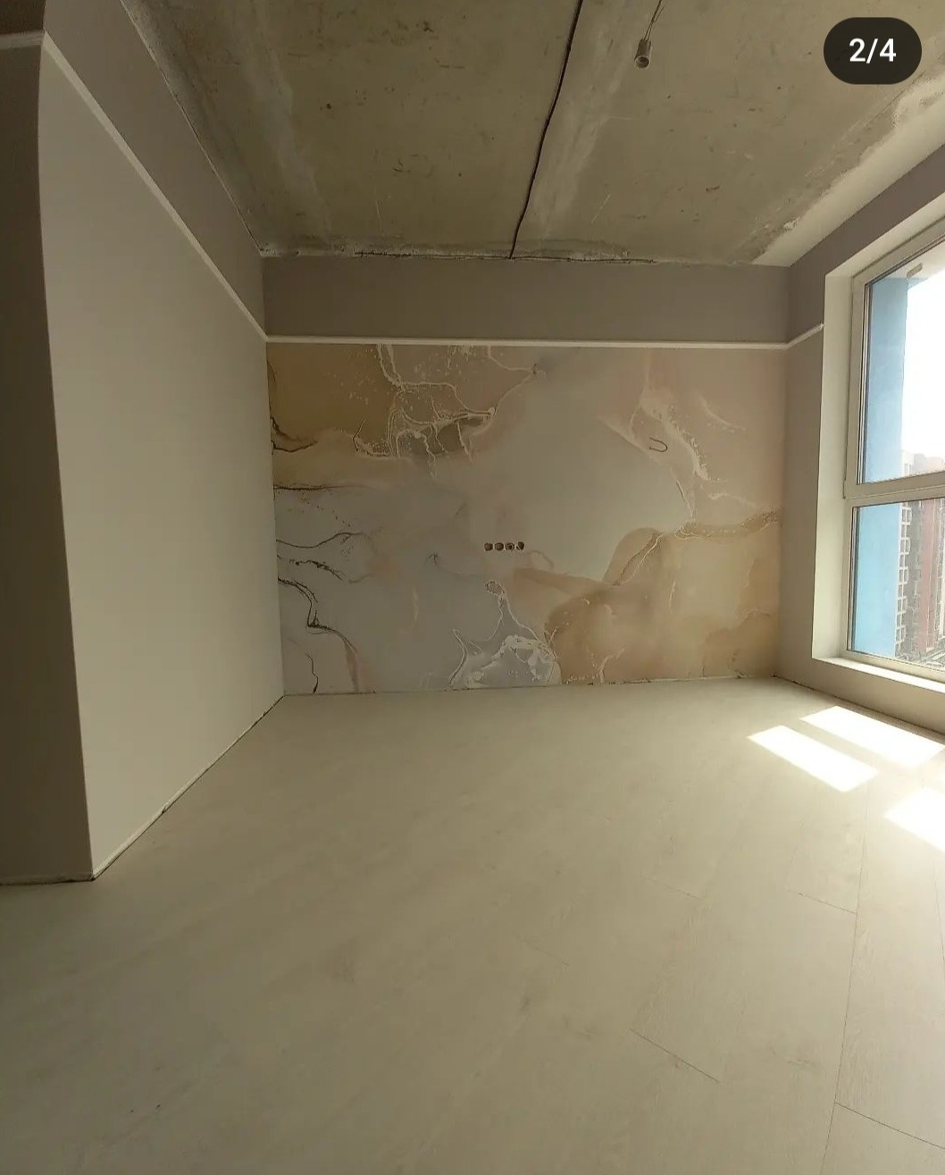 Wykończone wnętrze pokoju z beżową ścianą o marmurkowym wzorze, jasną podłogą i dużym oknem. Surowy betonowy sufit dodaje industrialnego charakteru.