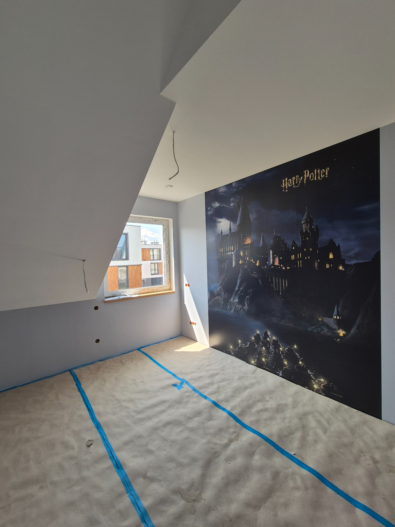 Pokój po remoncie z fototapetą Harry Potter. Podłoga zabezpieczona papierem i taśmą malarską. Ściany pomalowane na szaro, widoczne gniazdka. Okno z widokiem na budynek.