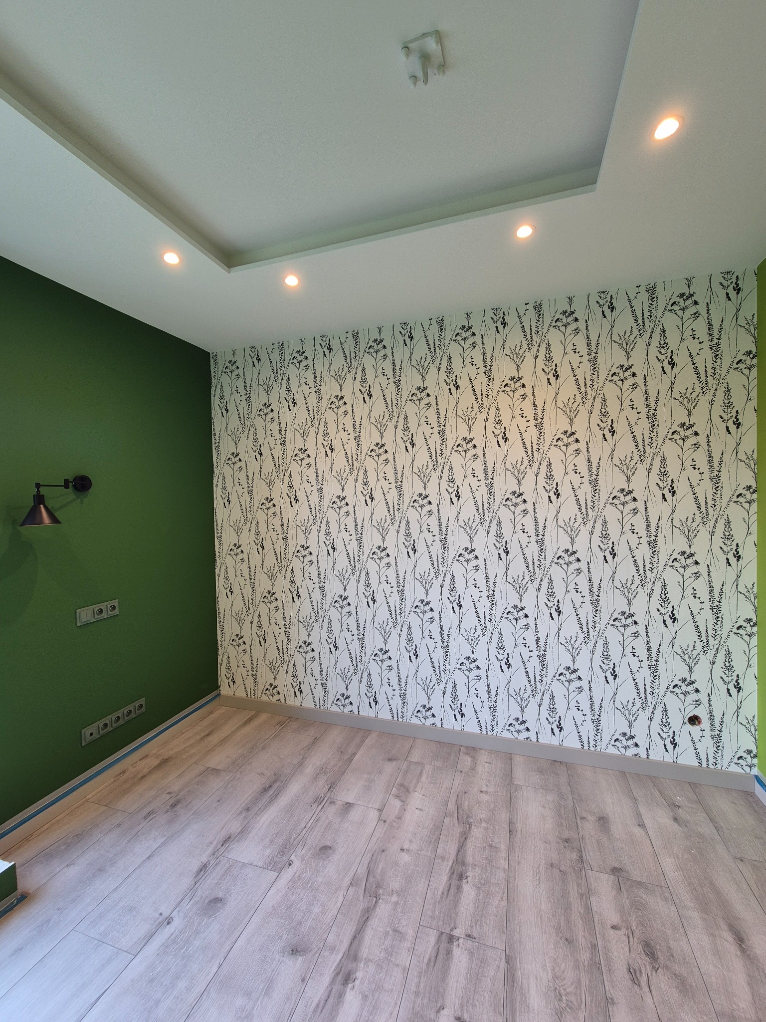 Ściana z tapetą w roślinny wzór, pomalowana na zielono ściana z lampą, panele podłogowe, sufit z oświetleniem punktowym. Wykończone wnętrze pokoju.