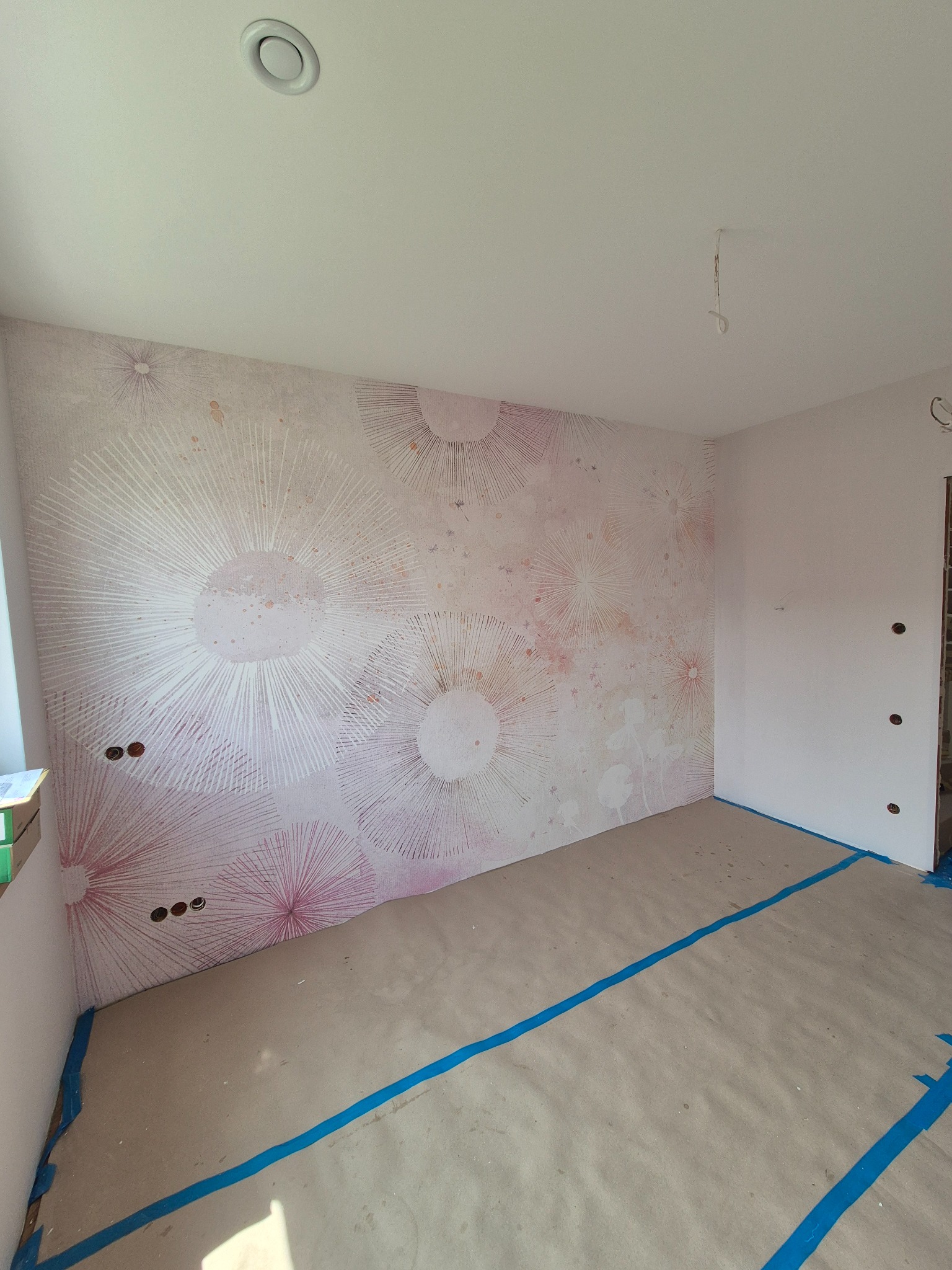 Pokój po tapetowaniu z wzorem w różowe kwiaty, podłoga zabezpieczona folią. Widoczne gniazdka elektryczne i sufit z wentylacją.