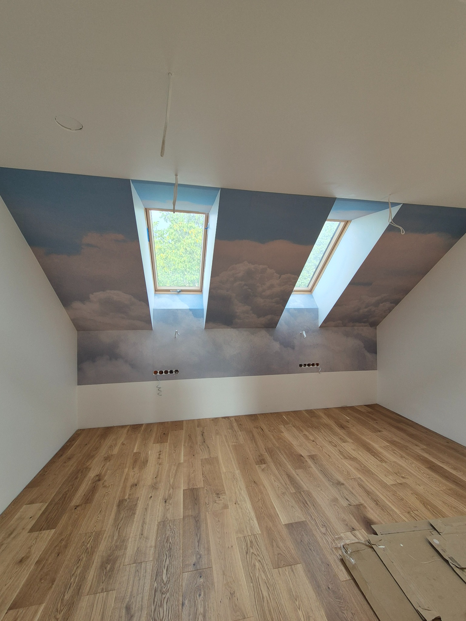 Pokój na poddaszu z drewnianą podłogą i tapetą z motywem chmur na skośnych ścianach. Dwa okna dachowe wpuszczają naturalne światło. Widoczne gniazdka elektryczne.