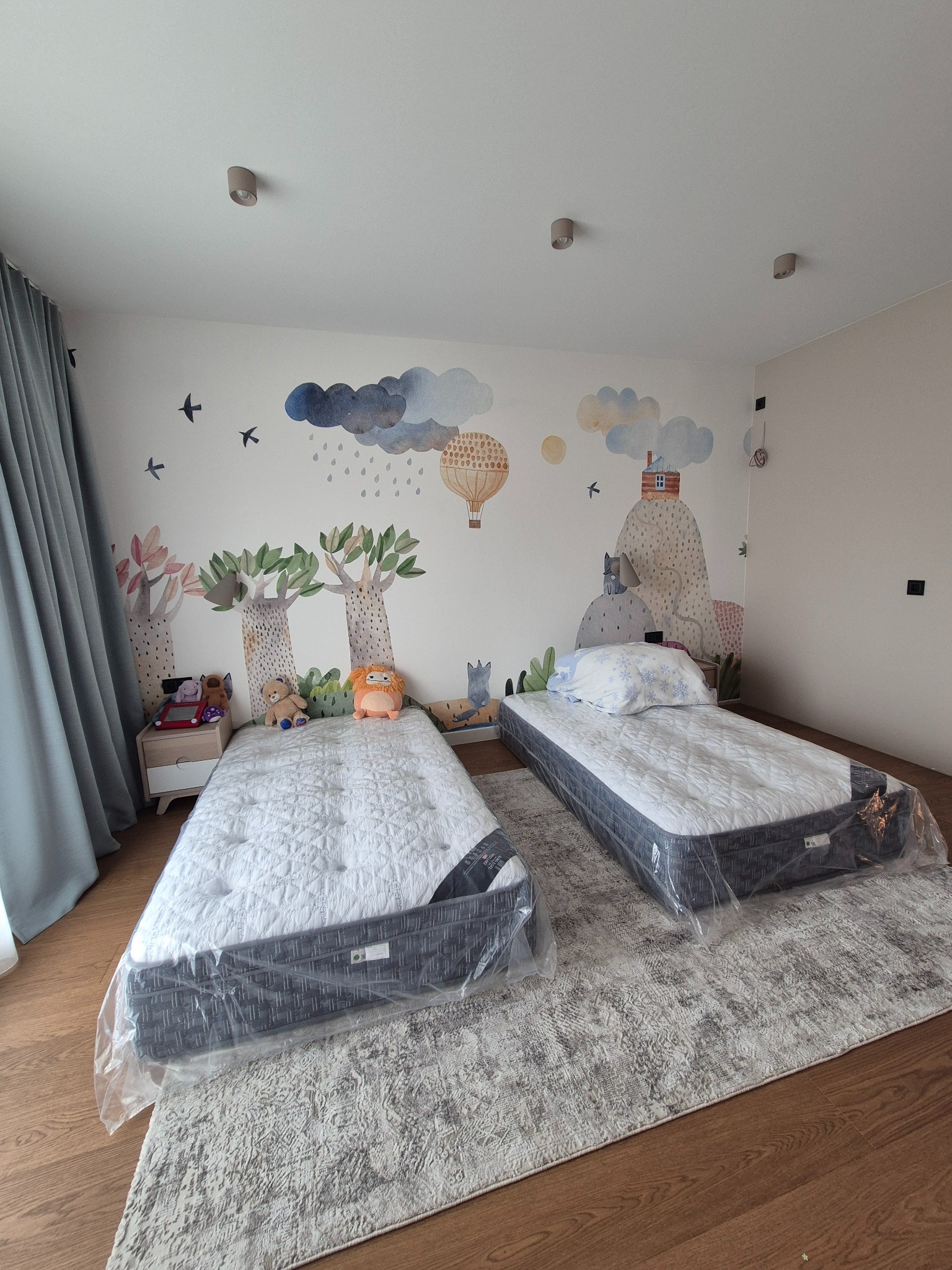 Dwa łóżka dziecięce w folii ochronnej, ustawione na dywanie w pokoju z malowidłami na ścianie przedstawiającymi drzewa i krajobraz.