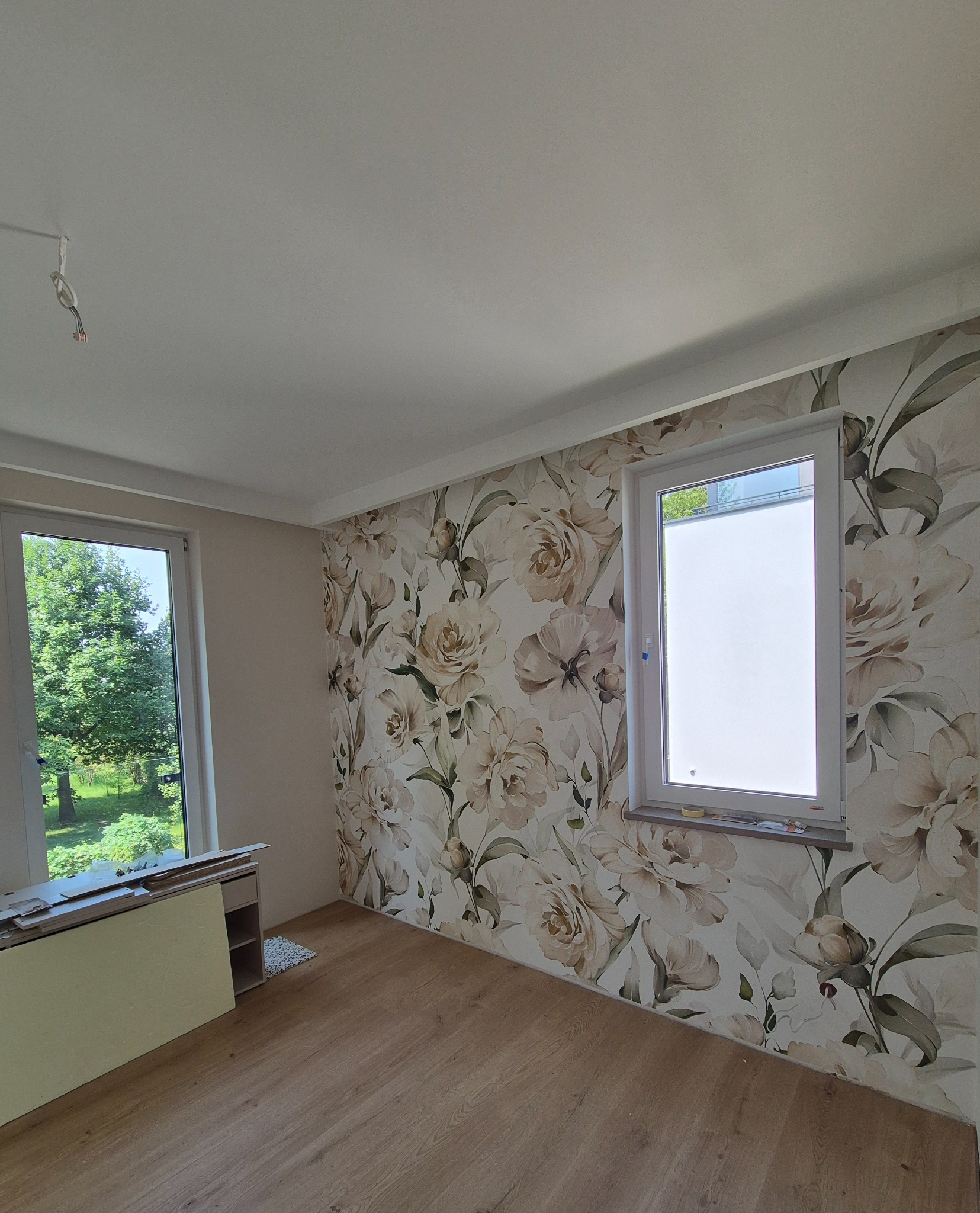 Pokój po tapetowaniu ścian z wzorem kwiatowym. Widoczne okna i drewniana podłoga. Częściowo widoczne materiały budowlane.
