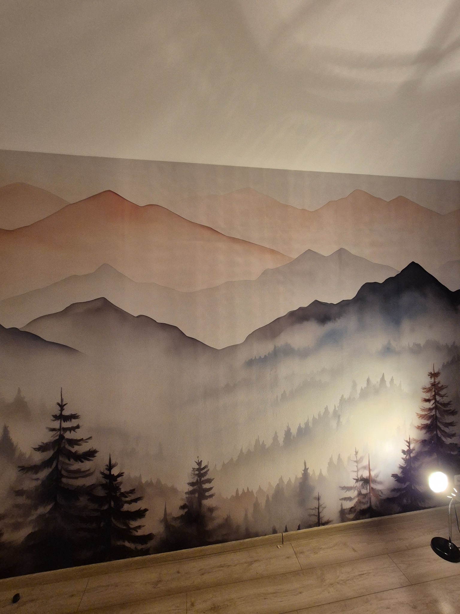Ściana z ręcznie malowanym pejzażem górskim w odcieniach brązu i szarości, z motywem lasu iglastego u dołu. Ciepłe światło lampki akcentuje fakturę malowidła.