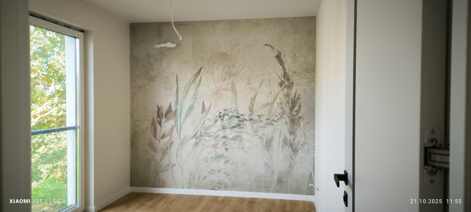 Pokój po tapetowaniu z motywem roślinnym na ścianie. Widoczne drewniane panele podłogowe, okno z widokiem na zieleń i fragment otwartych drzwi.