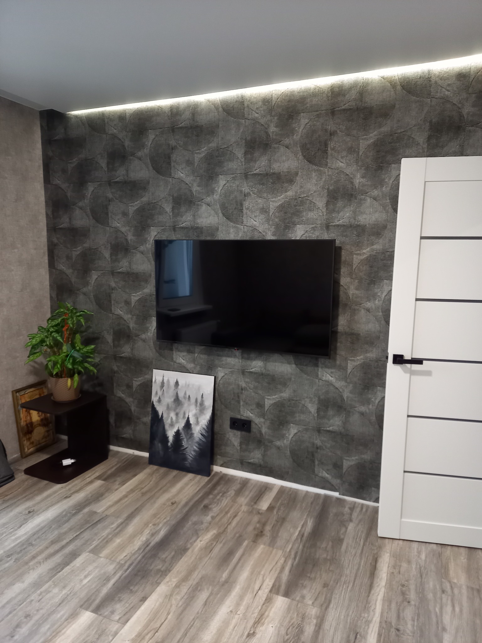 Nowoczesny salon z tapetą w geometryczne wzory, telewizorem na ścianie i drewnianą podłogą. Minimalistyczny wystrój z rośliną i obrazem leśnym.