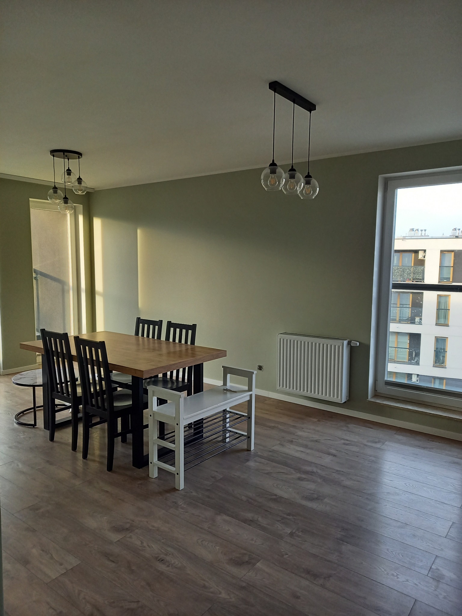 Jasne wnętrze pokoju z drewnianą podłogą i stołem z krzesłami. Na ścianie grzejnik i okno z widokiem na budynek. Dwie lampy sufitowe oświetlają przestrzeń.