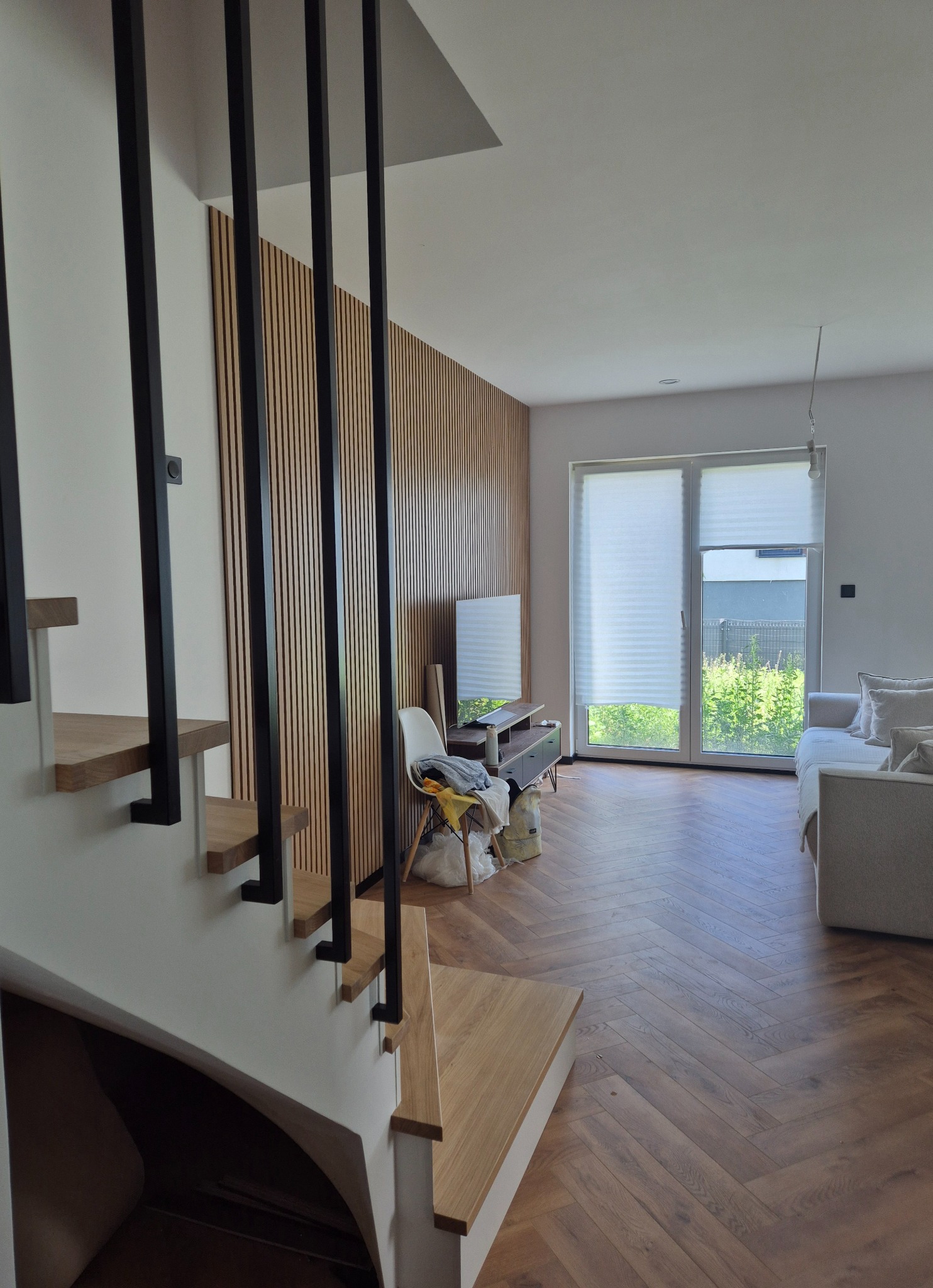 Nowoczesne wnętrze domu z drewnianą ścianą z lameli, schodami z drewnianymi stopniami i parkietem ułożonym w jodełkę. Widok na salon z oknem i sofą.
