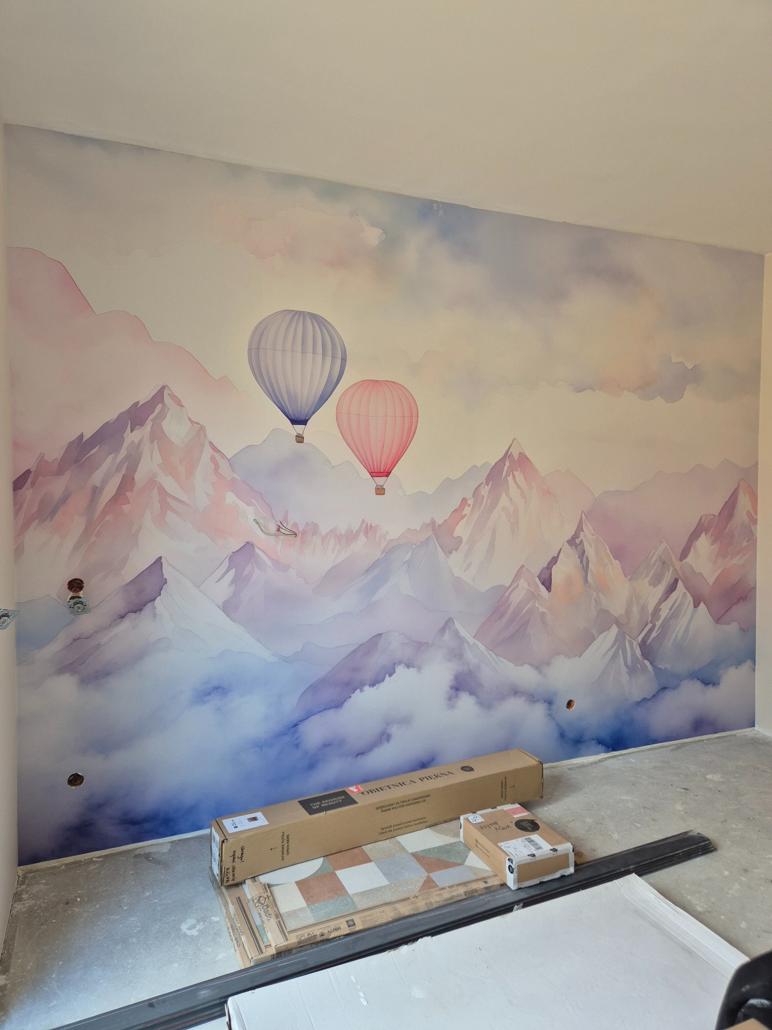Ściana z tapetą w pastelowe góry i balony, kartony z panelami podłogowymi u dołu. Widoczne gniazdka elektryczne. Ukończone tapetowanie pokoju.