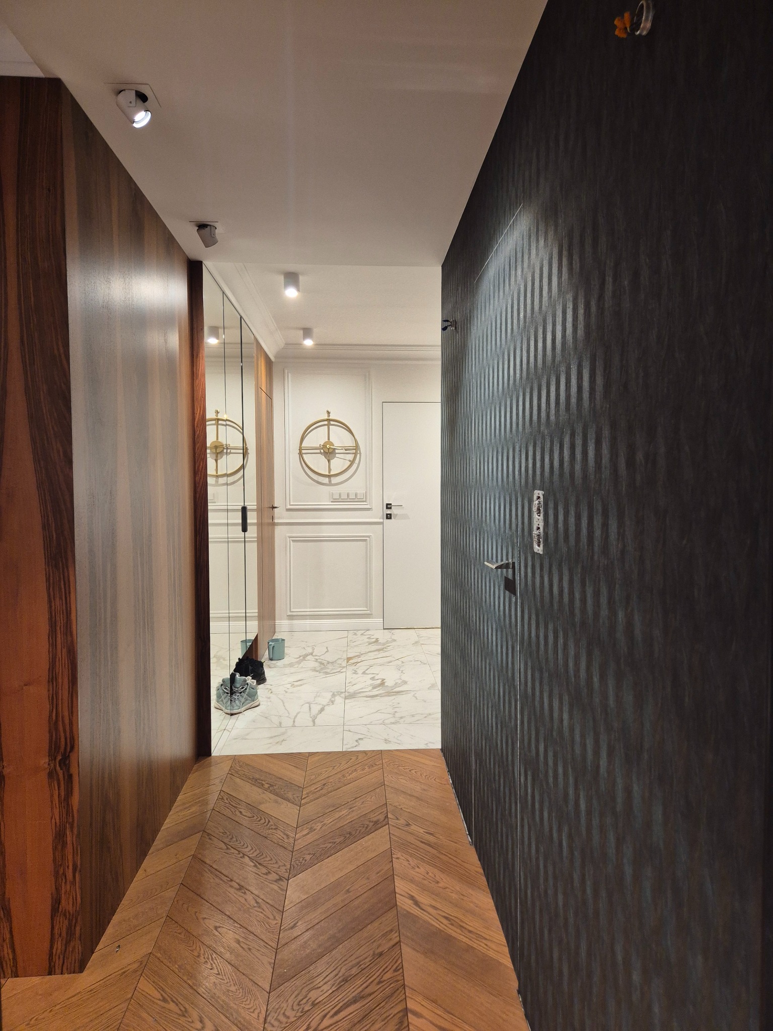 Elegancki korytarz po remoncie: parkiet w jodełkę, ciemna tapeta w pionowe pasy, lustro w zabudowie i marmurowe płytki w głębi. Nowoczesne oświetlenie.