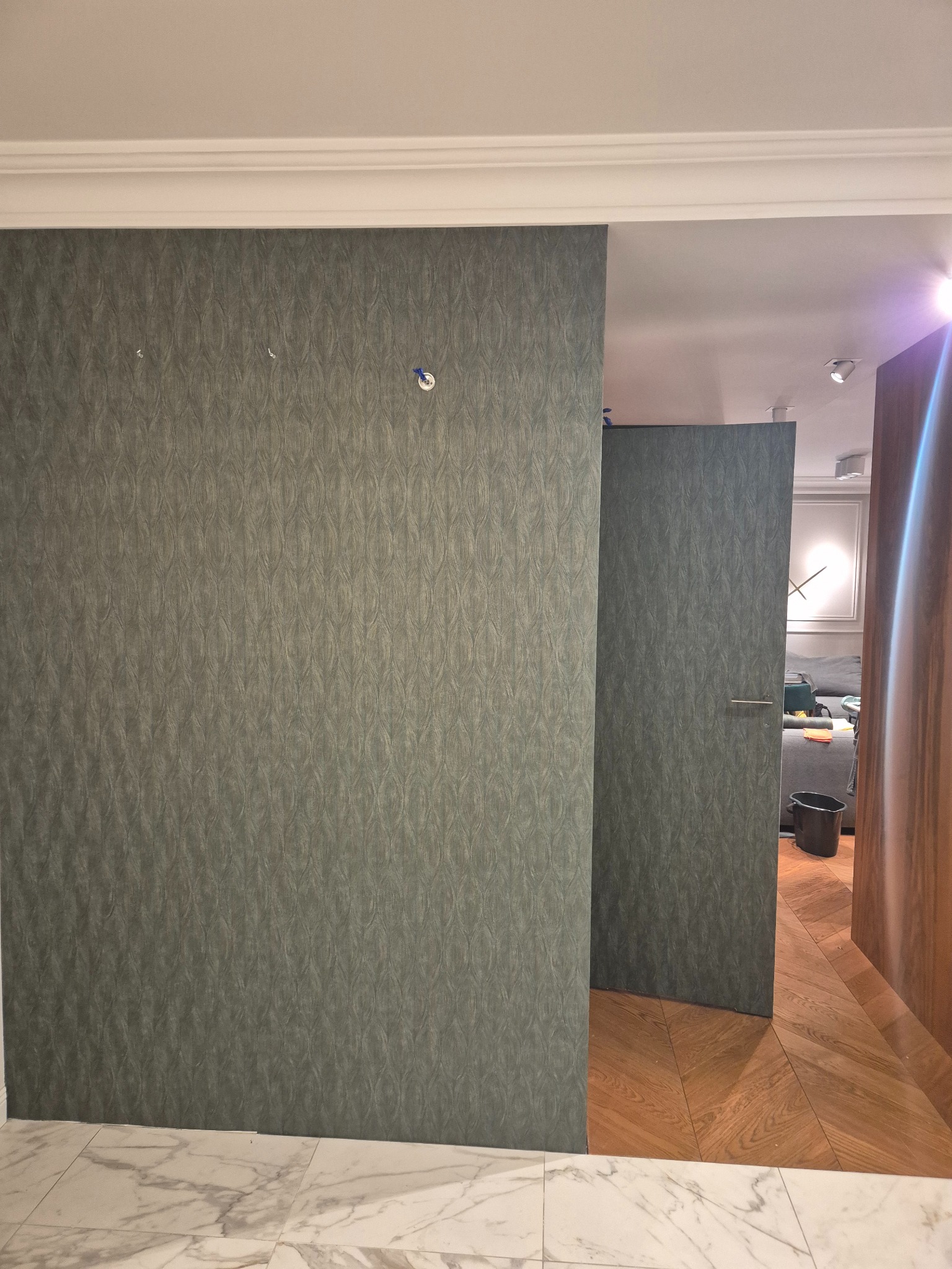 Tapeta w odcieniach szarości z subtelnym wzorem na ścianie, widoczna częściowo otwarta, dopasowana kolorystycznie przesuwna ściana działowa. Marmurowa podłoga i parkiet w jodełkę.