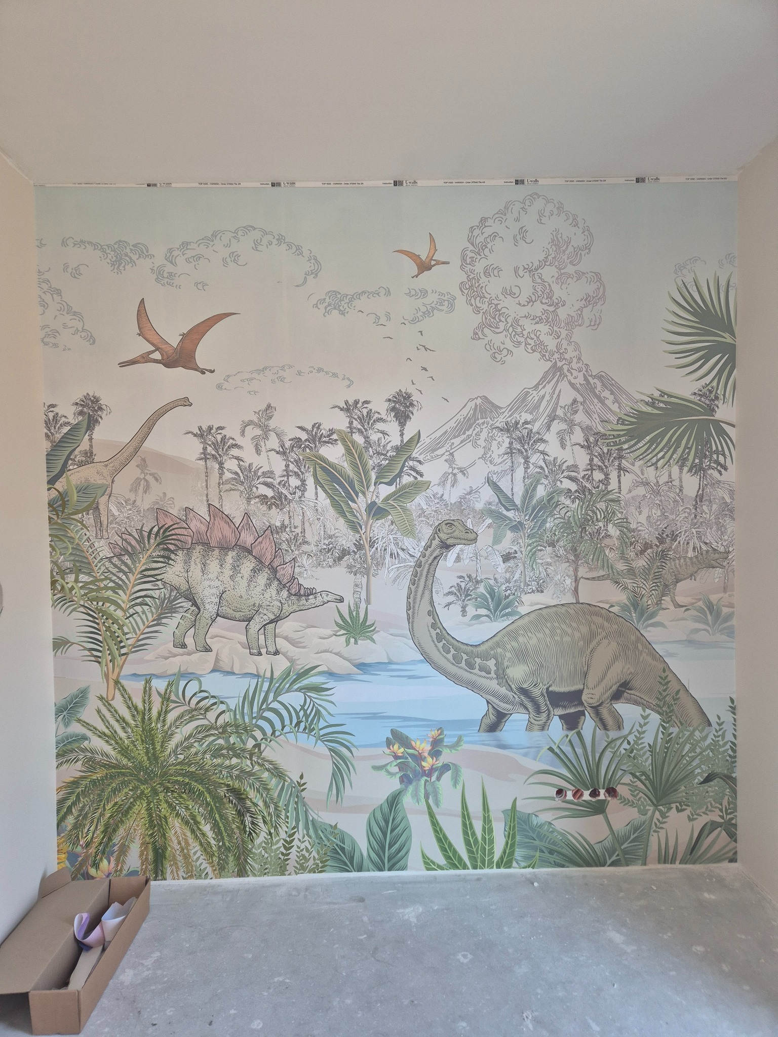 Ściana z tapetą w motywie prehistorycznego krajobrazu z dinozaurami i wulkanem, widoczne fragmenty rolek tapety w kartonie na podłodze.