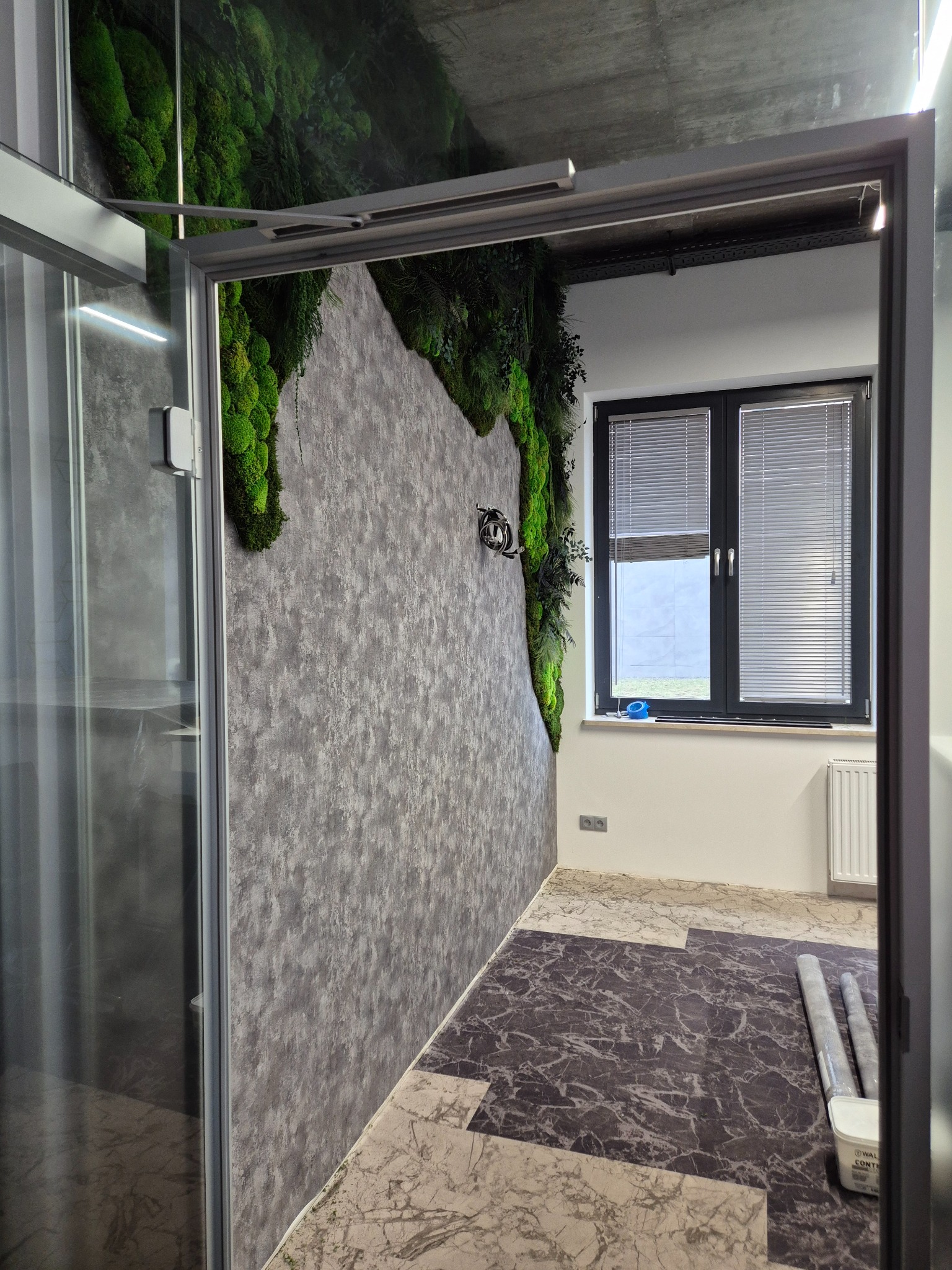 Nowoczesna łazienka z akcentem mchu na ścianie i wzorzystą podłogą. Widok przez otwarte drzwi kabiny prysznicowej. Wykończenie w szarościach i bieli, rolety w oknie.