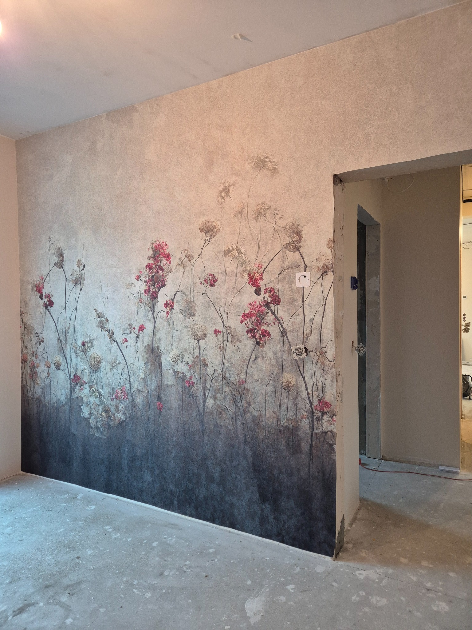 Ściana z fototapetą w kwiaty w odcieniach szarości i różu, widoczne gniazdka elektryczne i surowa podłoga. Ujęcie z niskiego kąta, widok na otwarte drzwi do innego pomieszczenia.