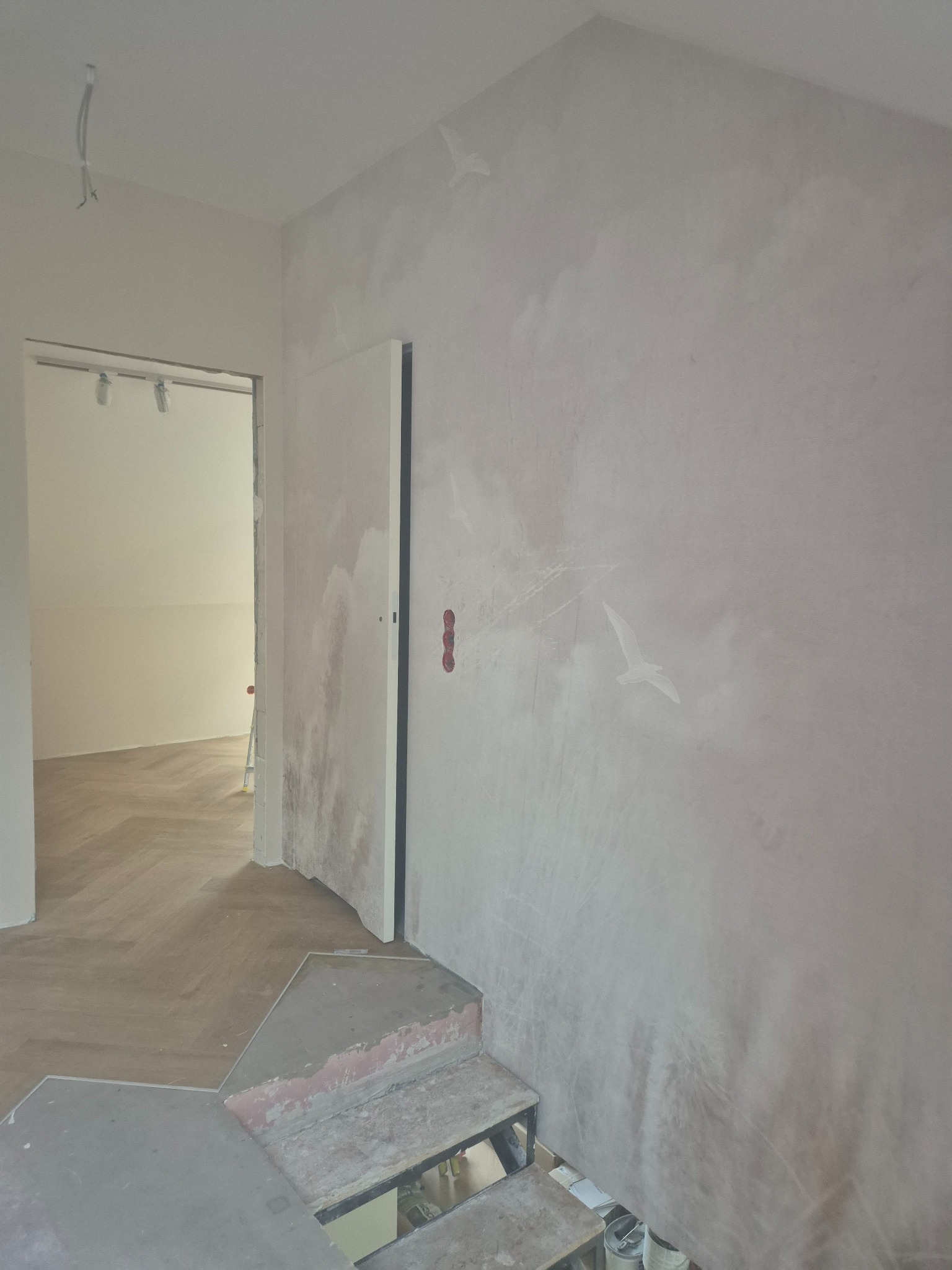 Ściana z dekoracyjnym malowaniem chmur i ptaków w jasnym wnętrzu. Widoczny fragment podłogi i schodów w trakcie prac wykończeniowych. Nietypowe wykończenie ściany.