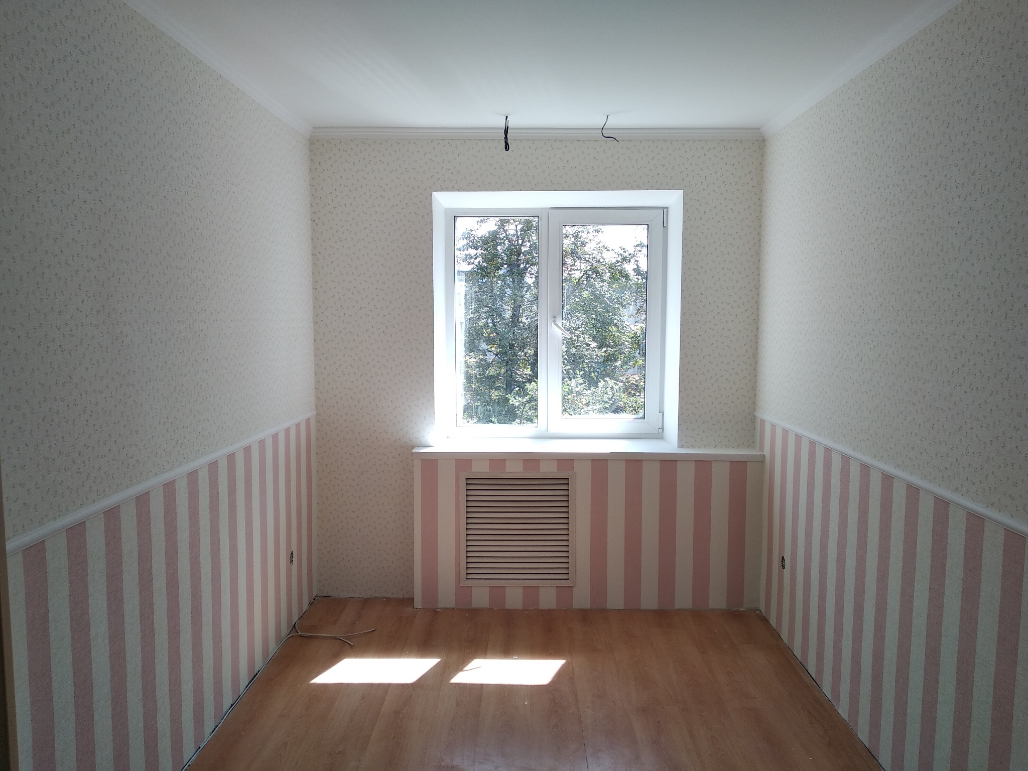 Pusty pokój po remoncie z tapetą w drobne wzory na górze i różowo-białymi pasami na dole, okno z widokiem na drzewa, drewniana podłoga i biały sufit z wystającymi kablami.