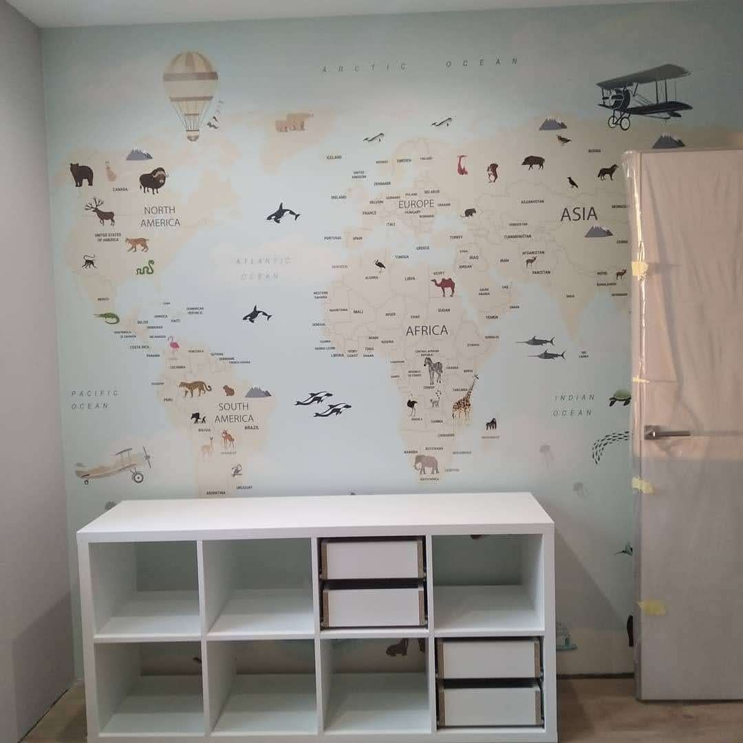 Ściana pokoju dziecięcego z tapetą w mapę świata z motywami zwierząt i pojazdów, biały regał z szufladami na pierwszym planie, drzwi z futryną po prawej stronie.