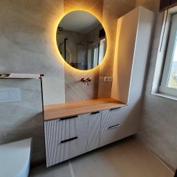 MEBS MARIUSZ SYC - Nowoczesna łazienka z szafką pod umywalkę z frezowanymi frontami, drewnianym blatem i okrągłym lustrem podświetlanym LED, widoczna toaleta w tle.