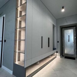 MEBS MARIUSZ SYC - Nowoczesna zabudowa przedpokoju z szafami na całej ścianie, półkami z podświetleniem LED i siedziskiem, utrzymana w szaro-białej kolorystyce z czarnymi uchwytami.