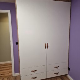 MEBS MARIUSZ SYC - Biała szafa dwudrzwiowa z czterema szufladami, z drewnianą ramą i uchwytami w kolorze brązowym, na tle fioletowej ściany i drewnianej podłogi, widoczny fragment otwartych drzwi do innego...