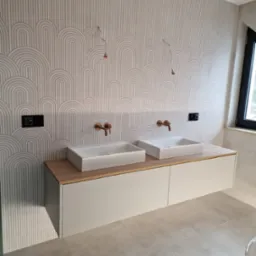 Dwie białe, prostokątne umywalki nablatowe z miedzianymi bateriami na szafce z jasnego drewna i białymi frontami, na tle ściany z dekoracyjną, geometryczną fakturą w odcieniach bieli i szarości.
