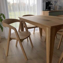 MEBS MARIUSZ SYC - Drewniany stół z krzesłami w jasnym wnętrzu. Widoczne detale wykonania mebli i wzorzysta ściana w tle. Naturalne światło podkreśla ciepłą kolorystykę drewna.