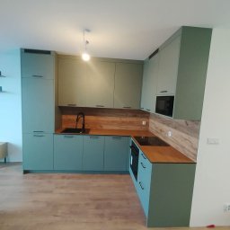 MEBS MARIUSZ SYC - Nowoczesna kuchnia w odcieniach zieleni z drewnianym blatem i czarną armaturą. Minimalistyczny design, fronty bez uchwytów, oświetlenie punktowe. Ujęcie z perspektywy podłogi.