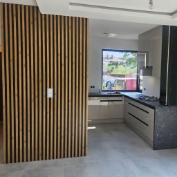 MEBS MARIUSZ SYC - Nowoczesna kuchnia z zabudową na wymiar, szare fronty, czarny blat i drewniana ścianka działowa z lameli. Minimalistyczny design, jasne wnętrze.