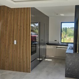 MEBS MARIUSZ SYC - Nowoczesna kuchnia z drewnianą ścianką działową i zabudową w odcieniach szarości. Minimalistyczny design, duże okno z widokiem na zieleń.