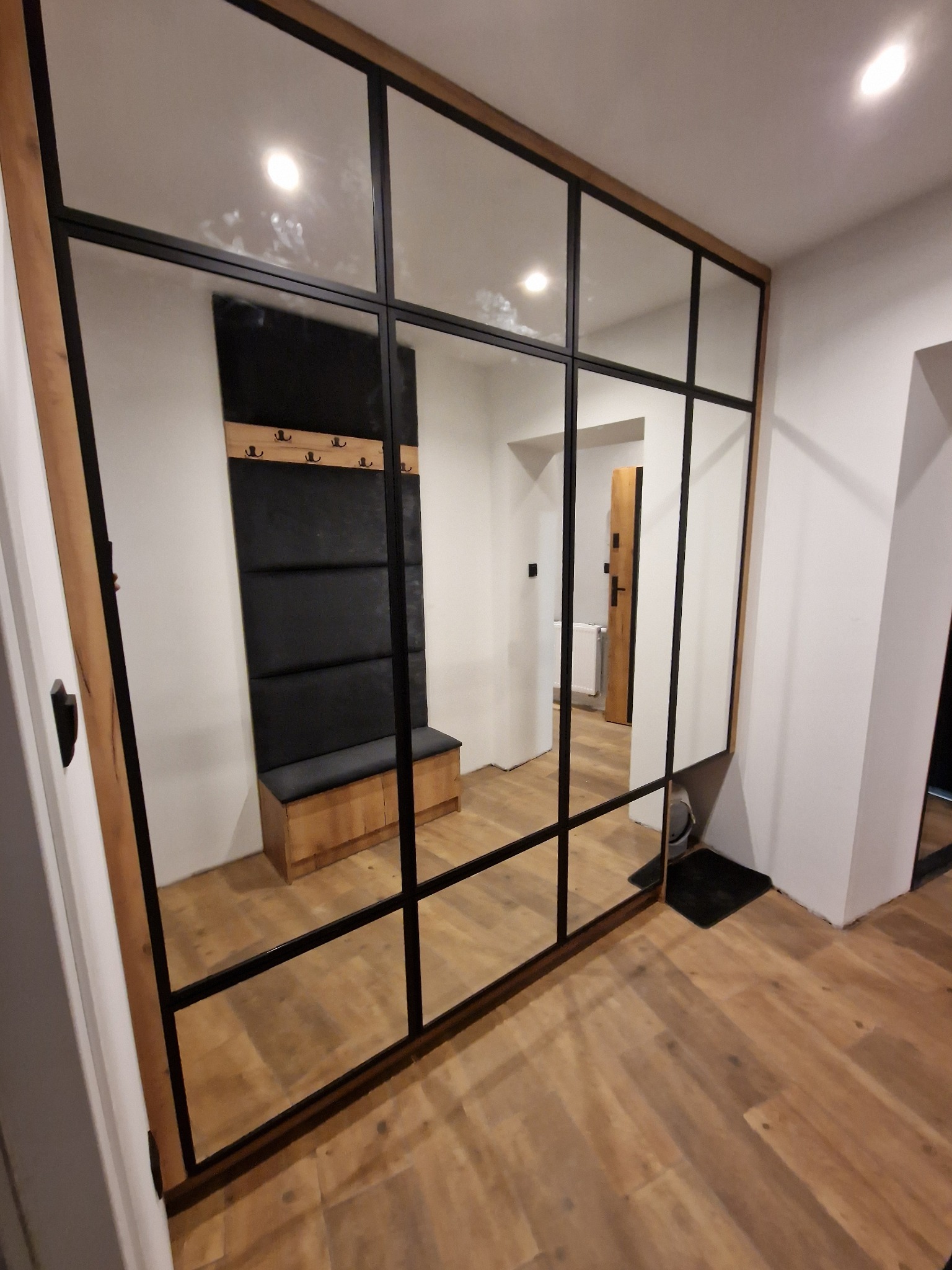 Przeszklona ściana działowa z czarnymi szprosami i drewnianą ramą, odbijająca wnętrze przedpokoju z siedziskiem, wieszakami i drewnianą podłogą.