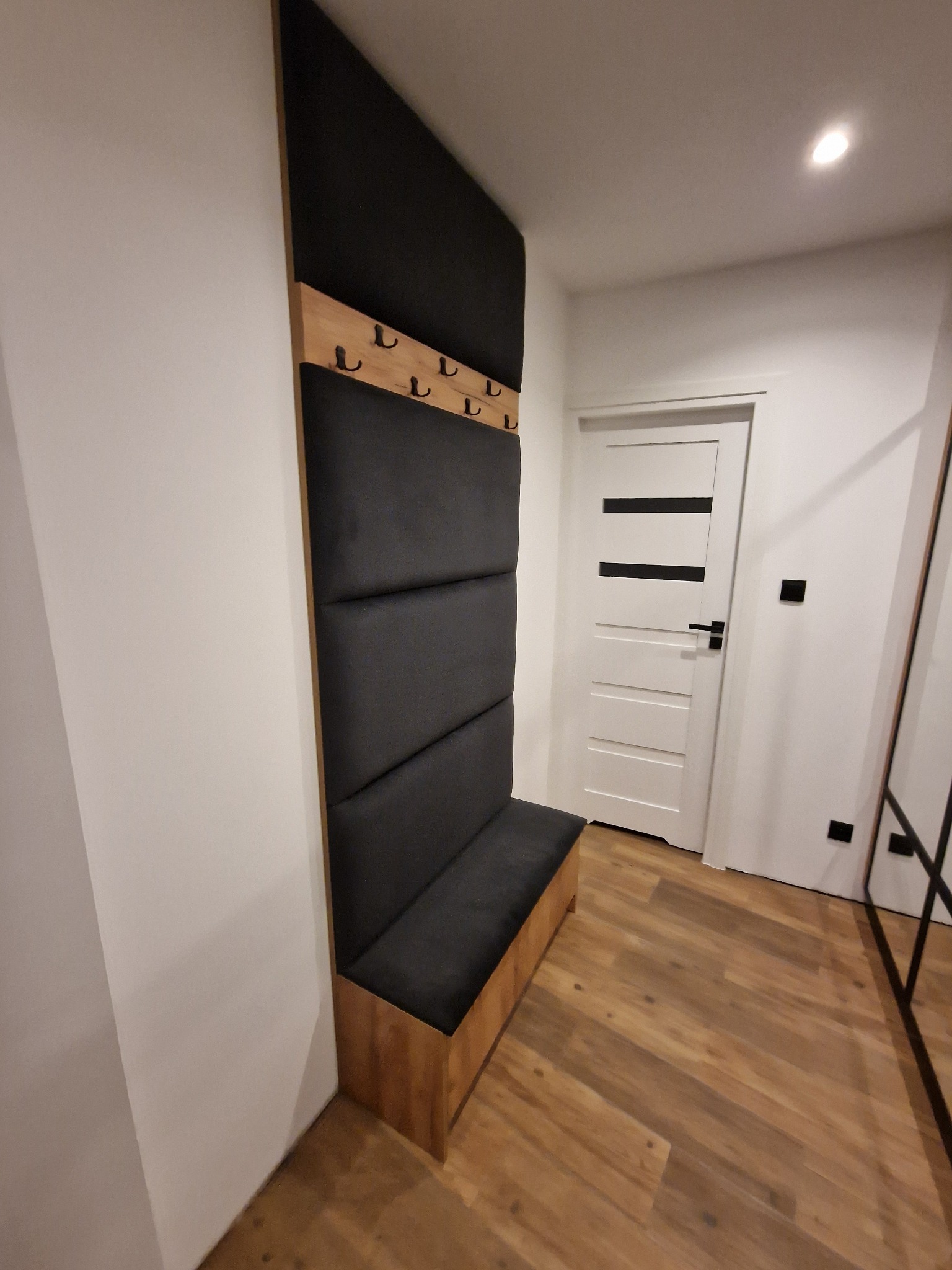 Panel ścienny z wieszakami i siedziskiem w przedpokoju, wykończony drewnem i czarną tapicerką, oświetlony punktowym światłem.