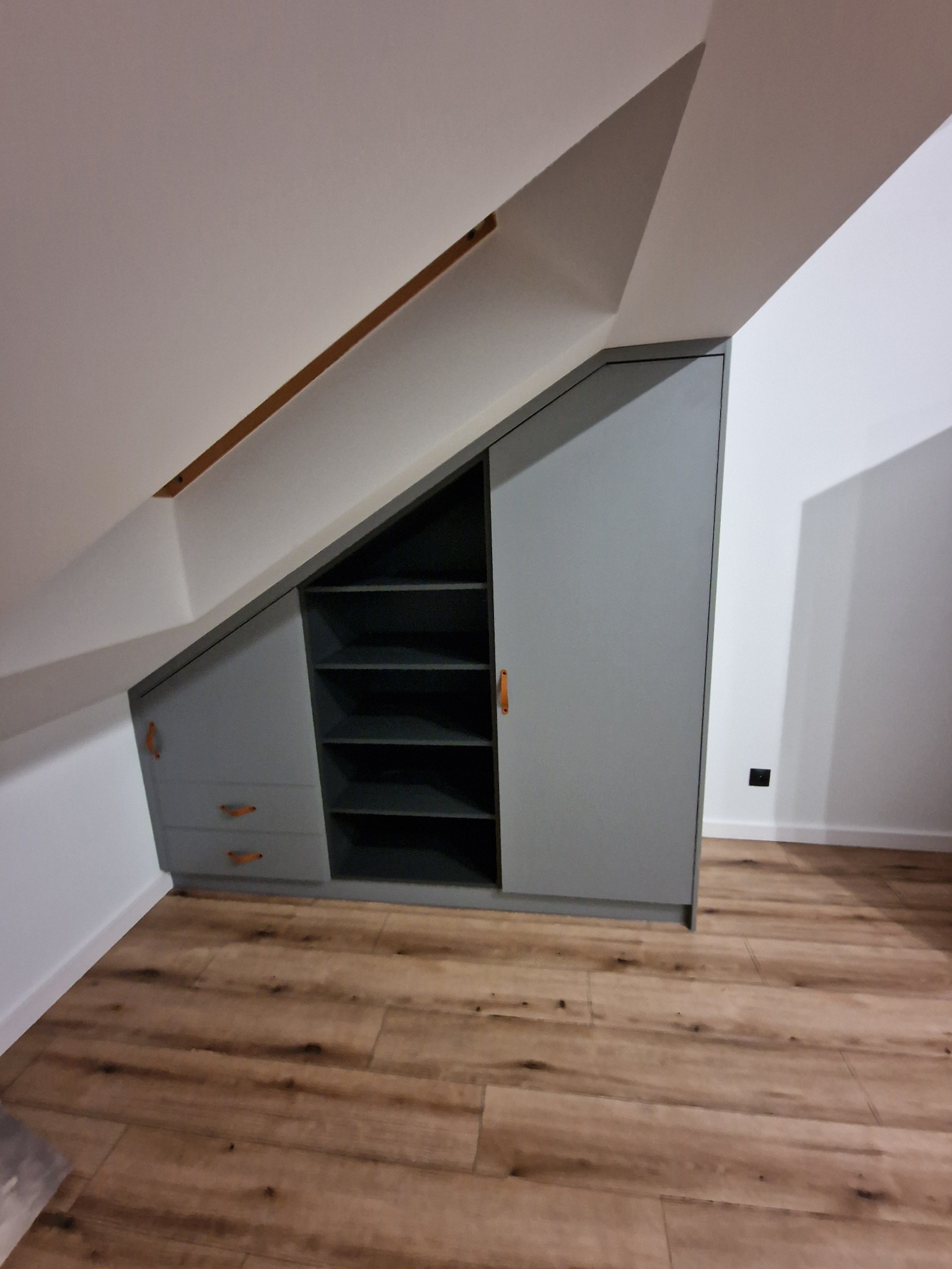 Szara zabudowa meblowa pod skosem na poddaszu z drewnianymi uchwytami, obejmująca szafę z drzwiami i otwartą półką.
