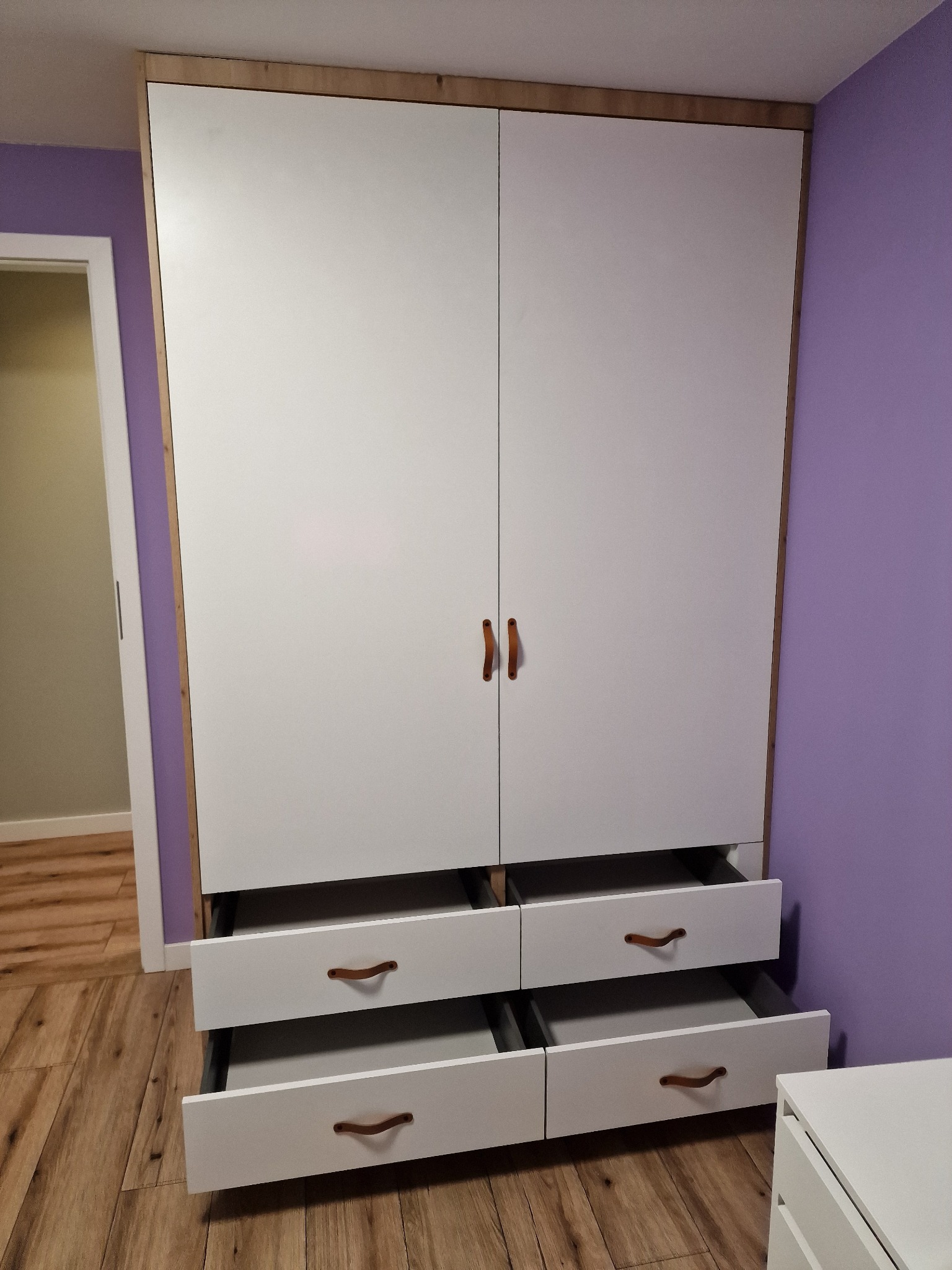 Biała szafa dwudrzwiowa z czterema wysuwanymi szufladami i drewnianymi uchwytami, umieszczona na tle fioletowej ściany i drewnianej podłogi, z otwartymi dwoma dolnymi szufladami.