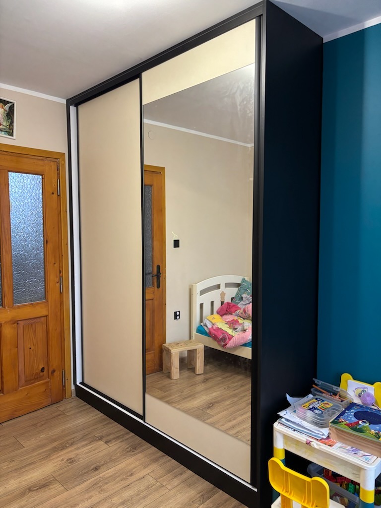 Szafa wnękowa z przesuwnymi drzwiami, beżowy front i lustro, czarne obramowanie, widoczny fragment pokoju dziecięcego w odbiciu lustrzanym.