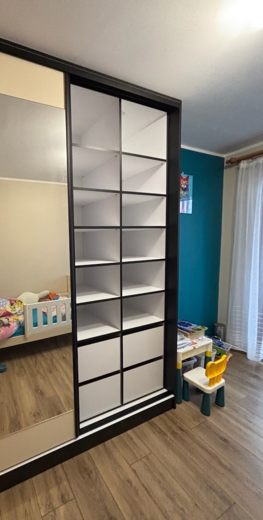 Wnętrze pokoju dziecięcego z dużą szafą przesuwną z lustrem i otwartymi półkami z czarnymi obramowaniami, widoczne łóżeczko i stolik z krzesełkiem.