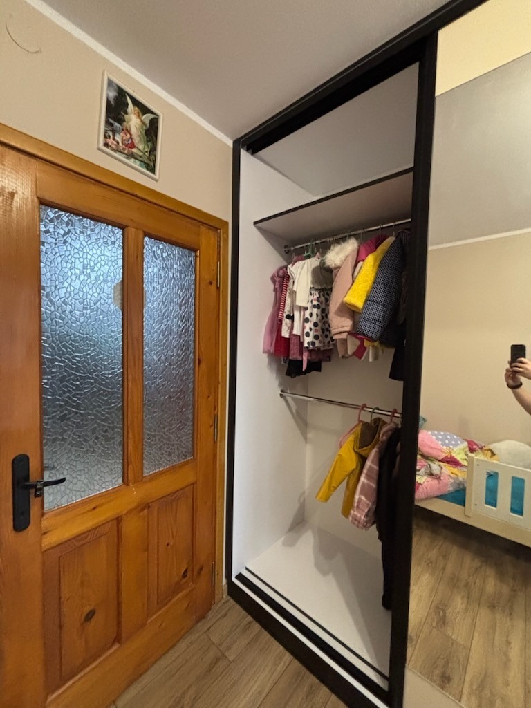 Wnętrze pokoju z drewnianymi drzwiami i otwartą szafą z przesuwnymi drzwiami, w której widać ubrania dziecięce na wieszakach; w lustrzanym odbiciu widoczne łóżko dziecięce.