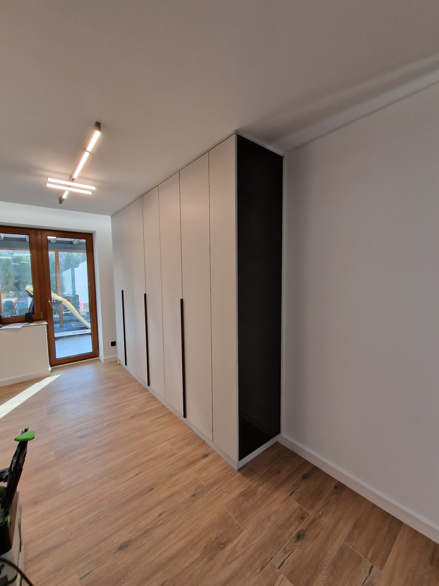 Biała szafa wnękowa z czarnymi, minimalistycznymi uchwytami, z otwartą czarną wnęką na końcu, na podłodze z paneli imitujących drewno, widok z perspektywy pomieszczenia.