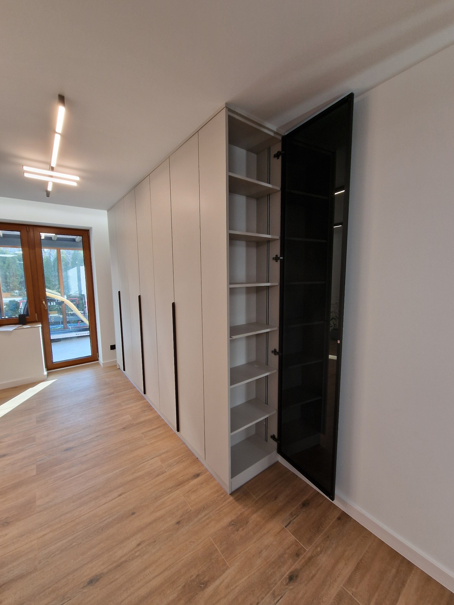 Biała szafa wnękowa z czarnymi, długimi uchwytami i otwartymi drzwiami z czarnego szkła, ukazującymi jasne półki wewnątrz.