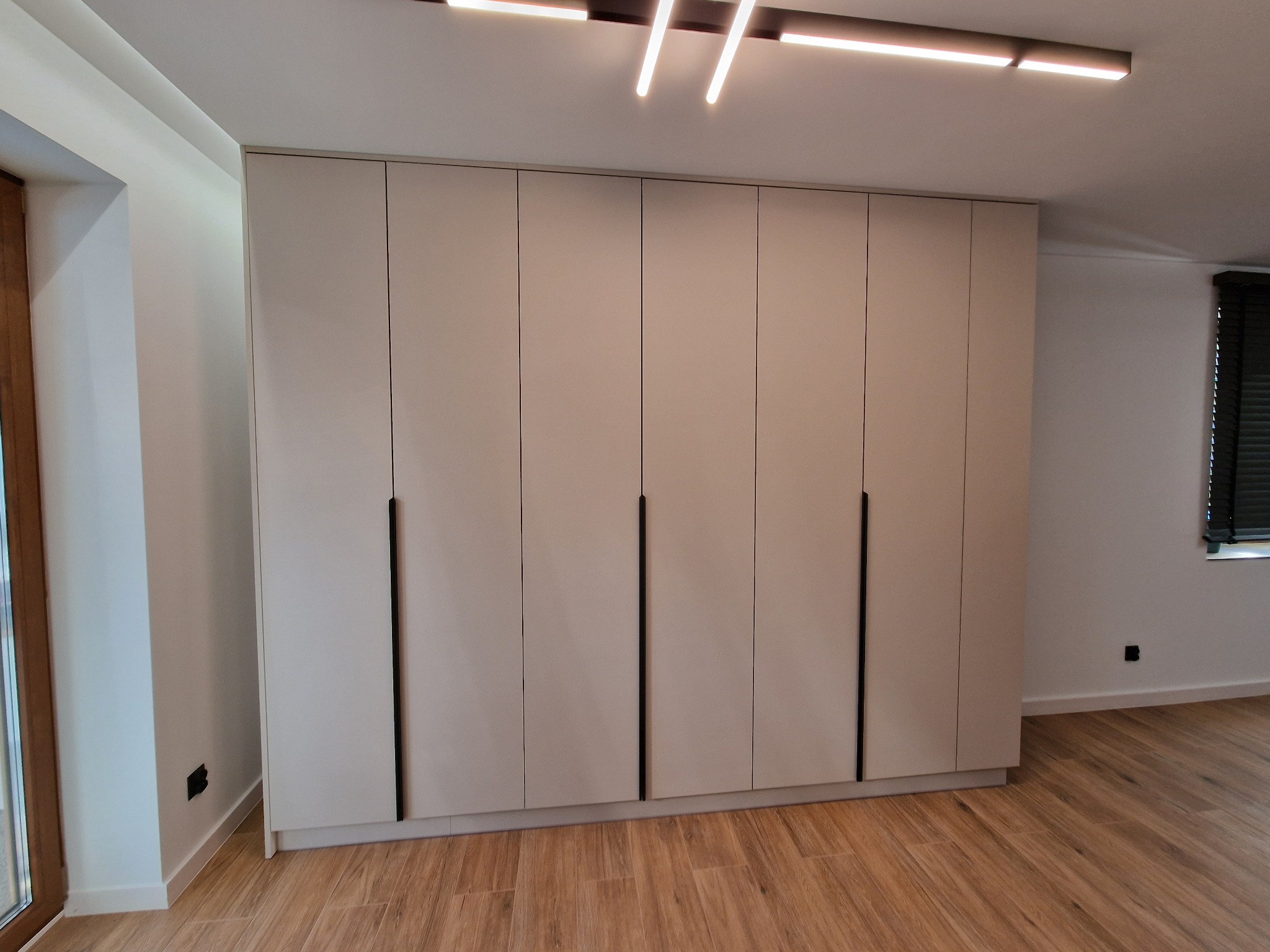 Duża, jasna szafa z sześcioma drzwiami i długimi, pionowymi uchwytami na tle drewnianej podłogi i nowoczesnego oświetlenia sufitowego.