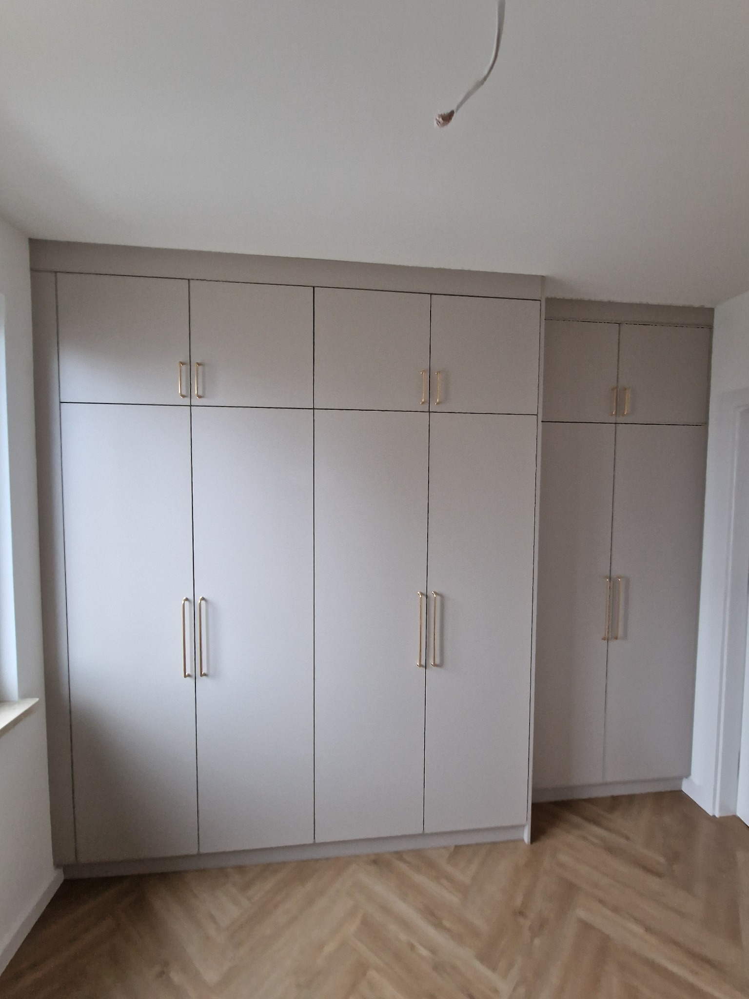 Jasnoszare szafy w zabudowie z podłużnymi złotymi uchwytami na tle parkietu ułożonego w jodełkę i białych ścian.