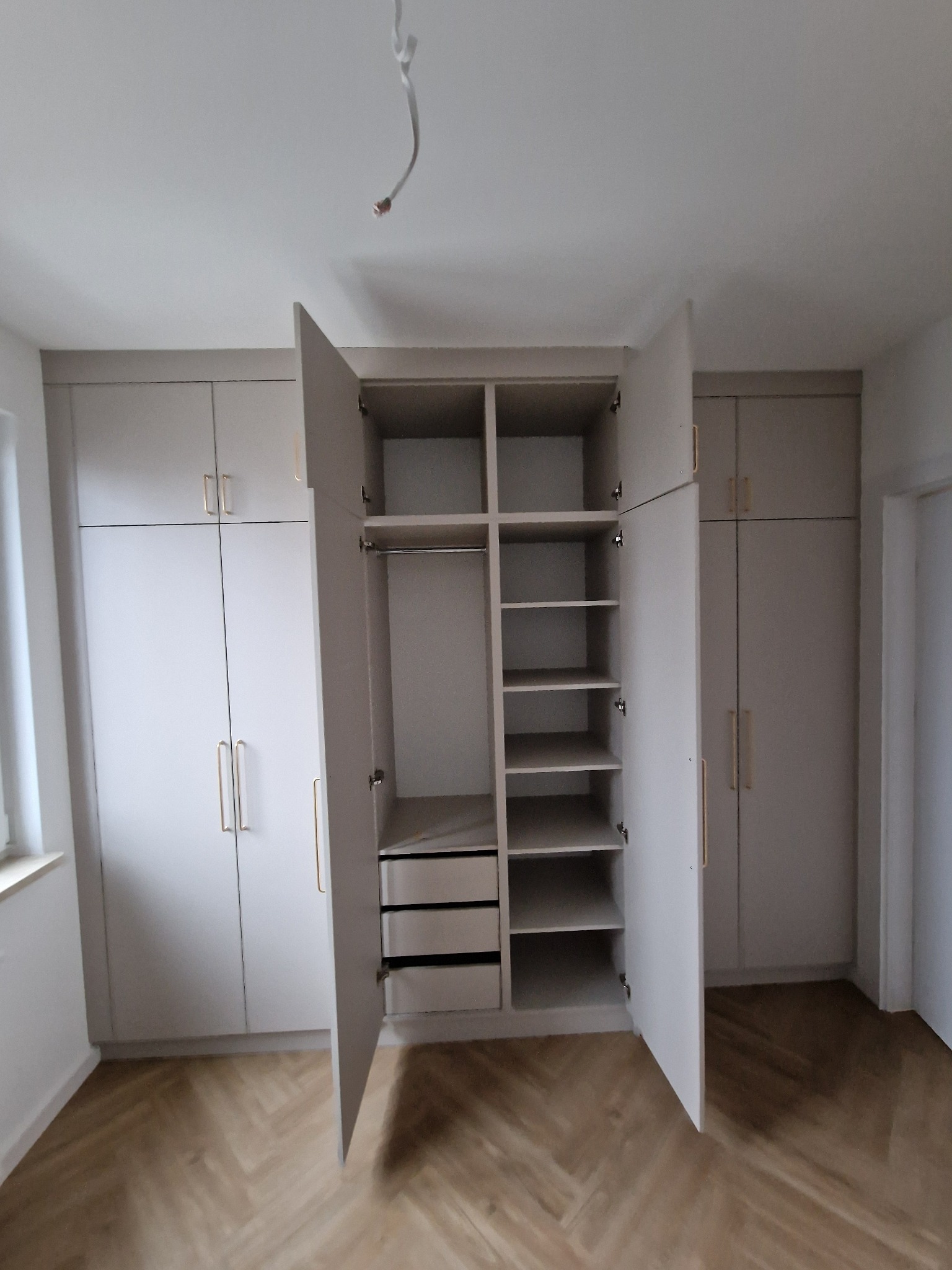 Jasna zabudowa meblowa z otwartymi drzwiami szafy, ukazująca półki, szuflady i drążek na ubrania, na tle parkietu ułożonego w jodełkę.