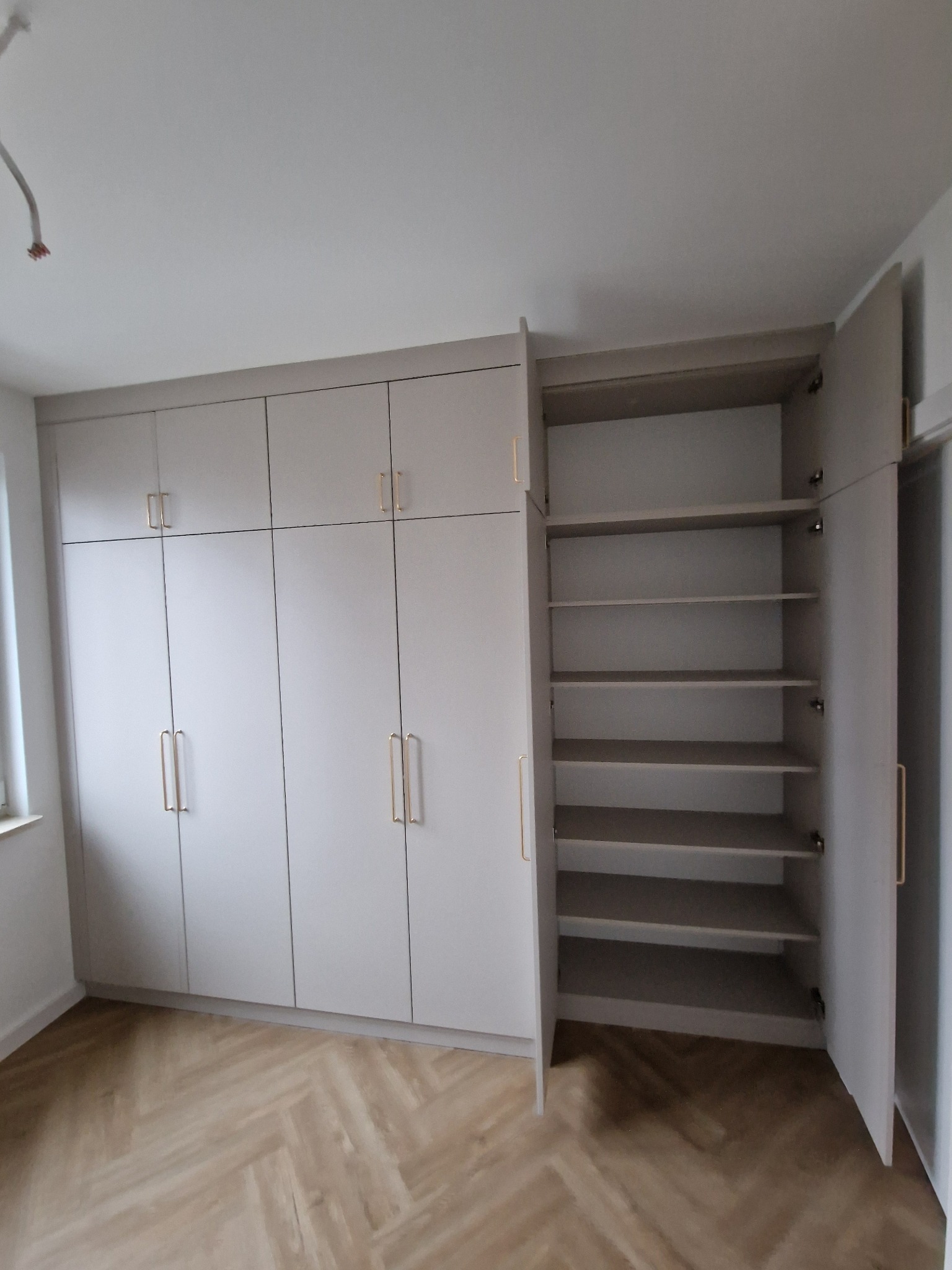 Jasnoszara zabudowa meblowa od podłogi do sufitu z złotymi uchwytami, jedna szafa otwarta ukazująca półki, parkiet ułożony w jodełkę.