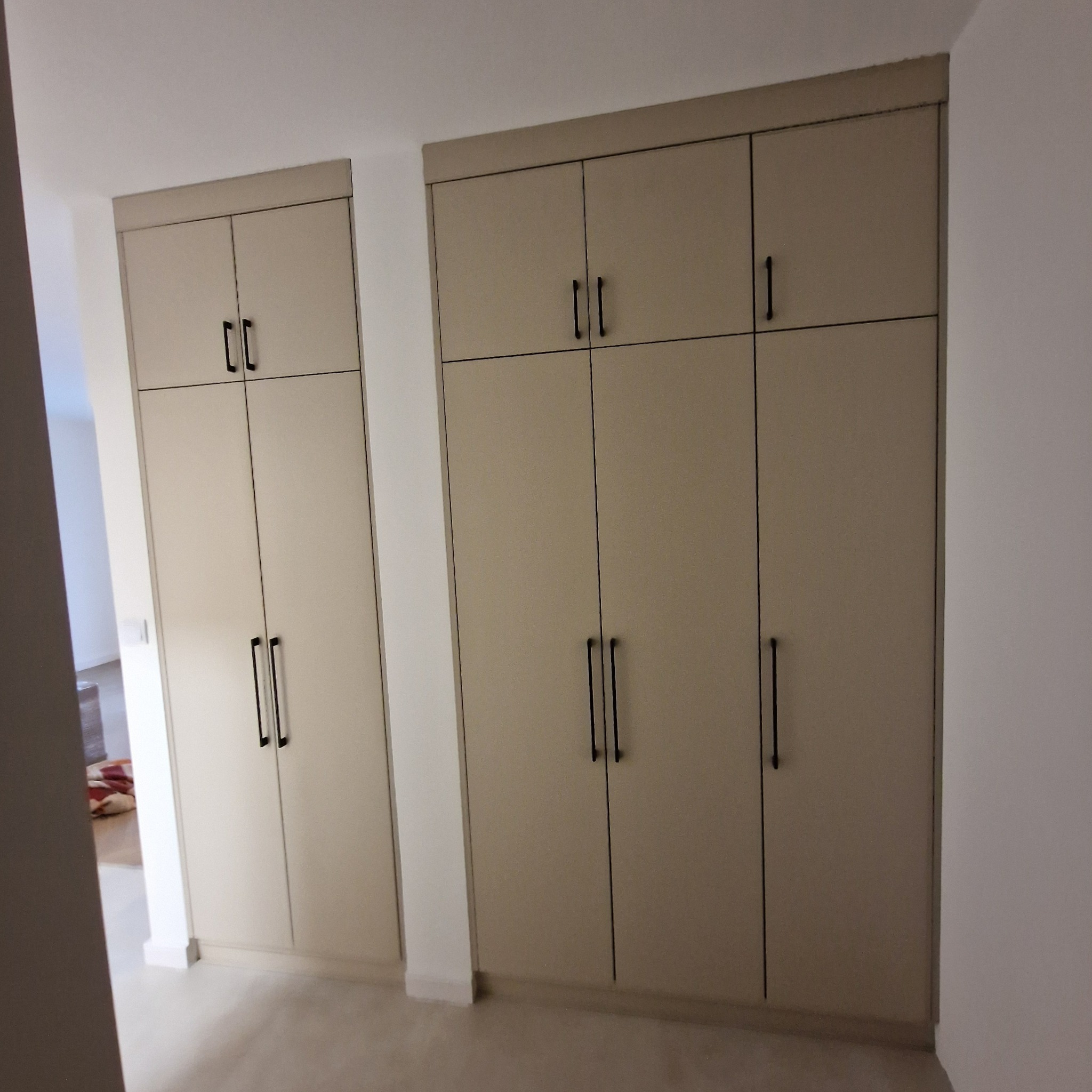 Dwie szafy wnękowe w odcieniach beżu z czarnymi, minimalistycznymi uchwytami, umieszczone w jasnym pomieszczeniu z widocznym fragmentem innego pokoju w tle.