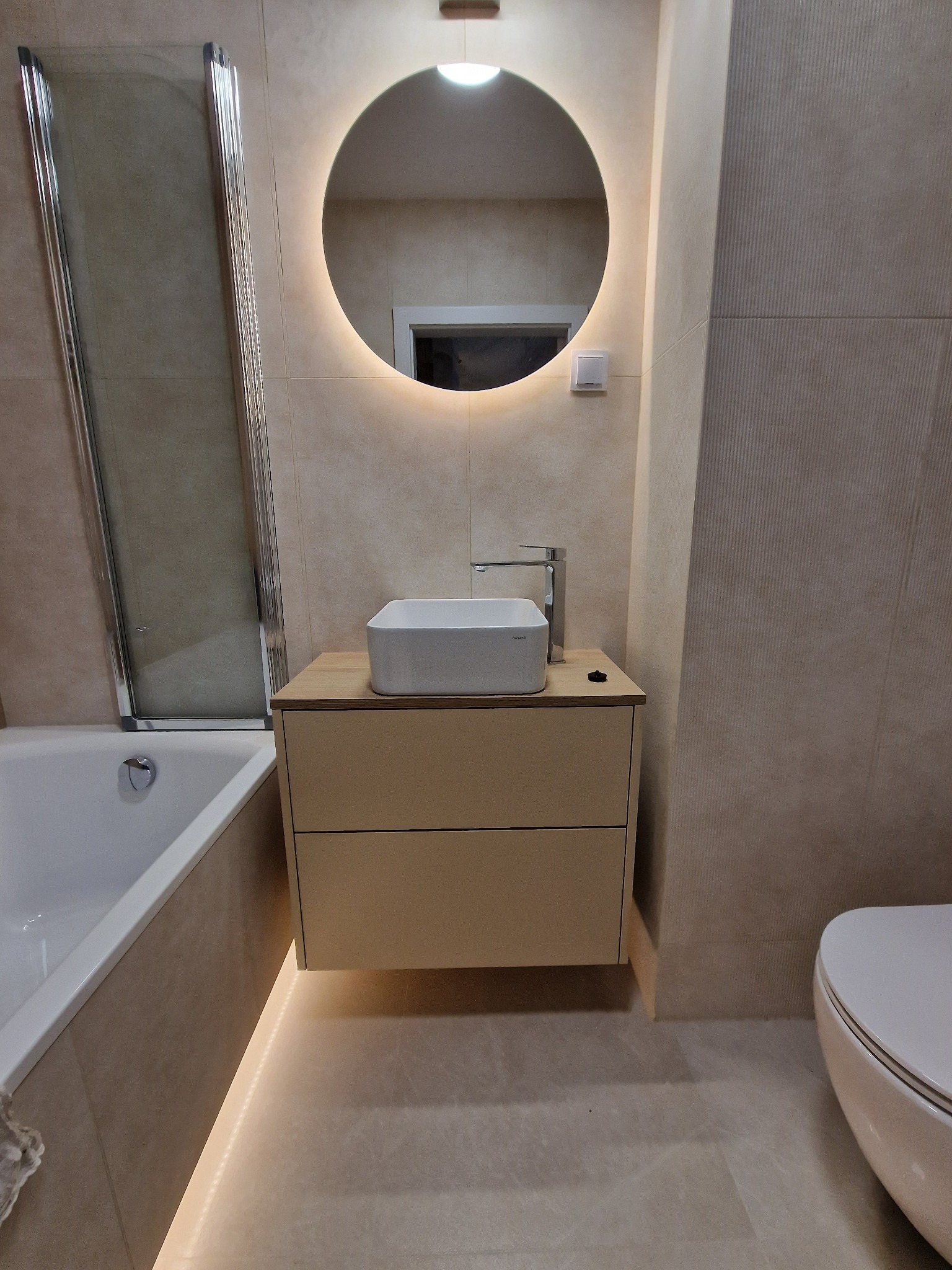 Nowoczesna łazienka z podświetlanym lustrem, umywalką nablatową na szafce z dwiema szufladami i podświetleniem pod szafką, obok wanna z parawanem nawannowym.