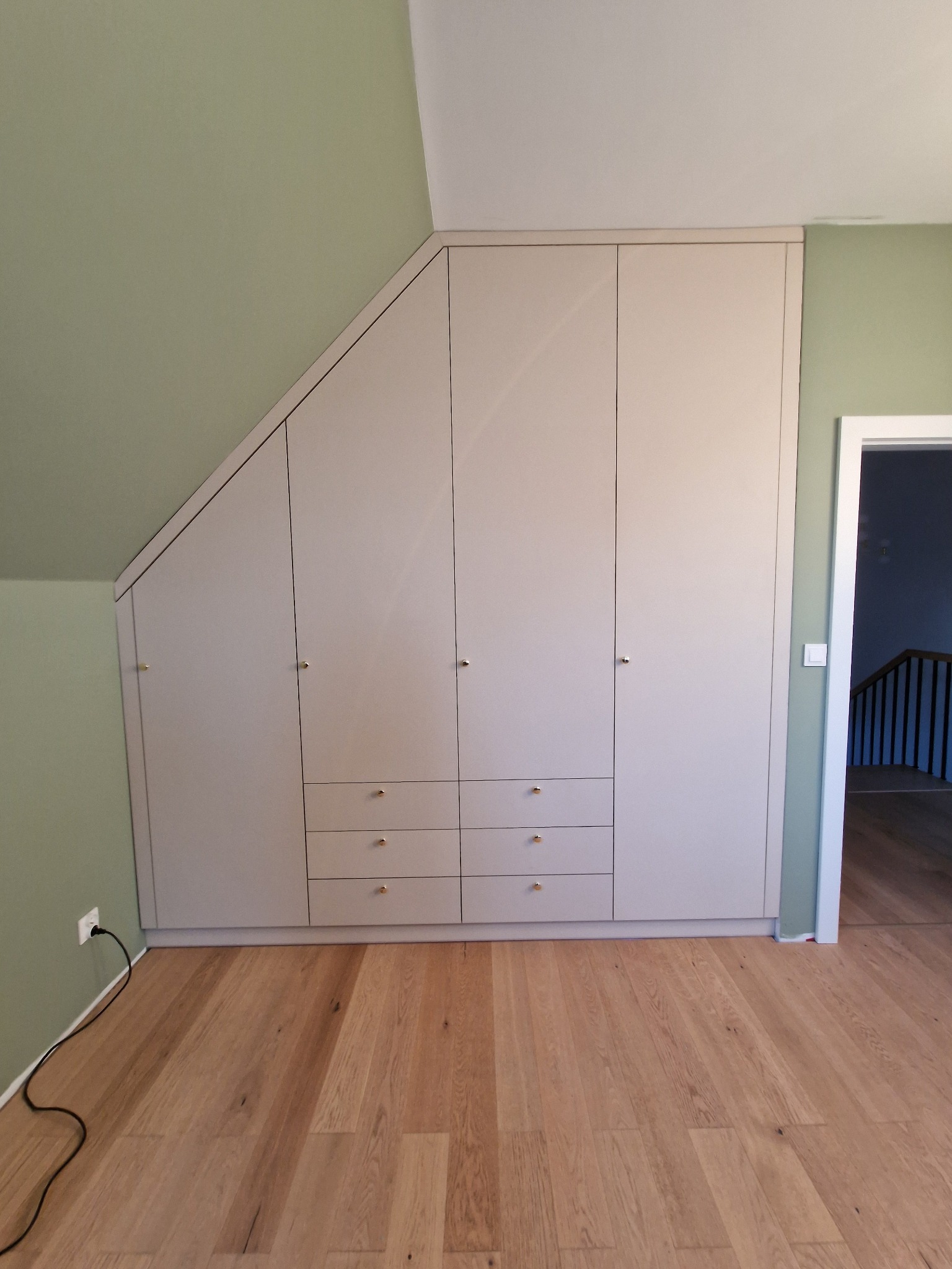 Jasna zabudowa meblowa pod skosem z szufladami i złotymi gałkami na tle zielonej ściany i drewnianej podłogi.