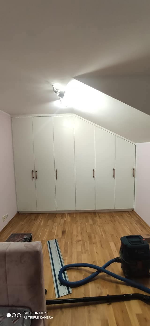 Biała zabudowa meblowa pod skosem na poddaszu, z drewnianymi uchwytami, na podłodze widoczne narzędzia i odkurzacz.