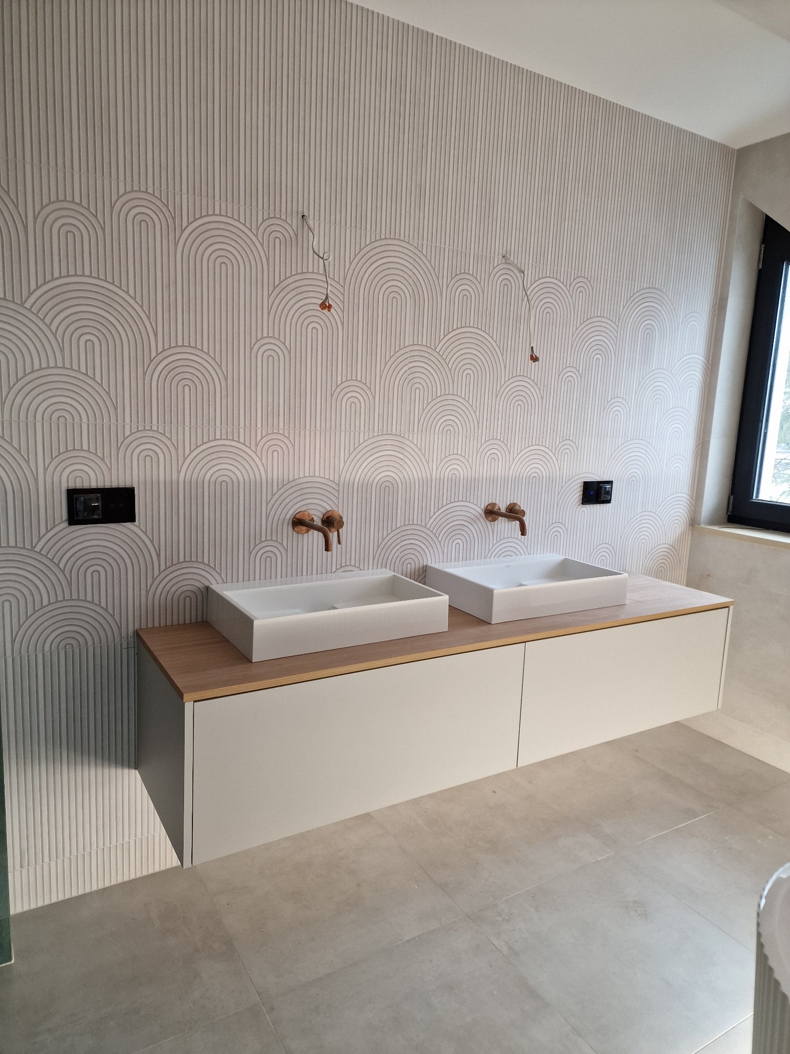 Dwie białe, prostokątne umywalki nablatowe z miedzianymi bateriami na szafce z jasnego drewna i białymi frontami, na tle ściany z dekoracyjną, geometryczną fakturą w odcieniach bieli i szarości.