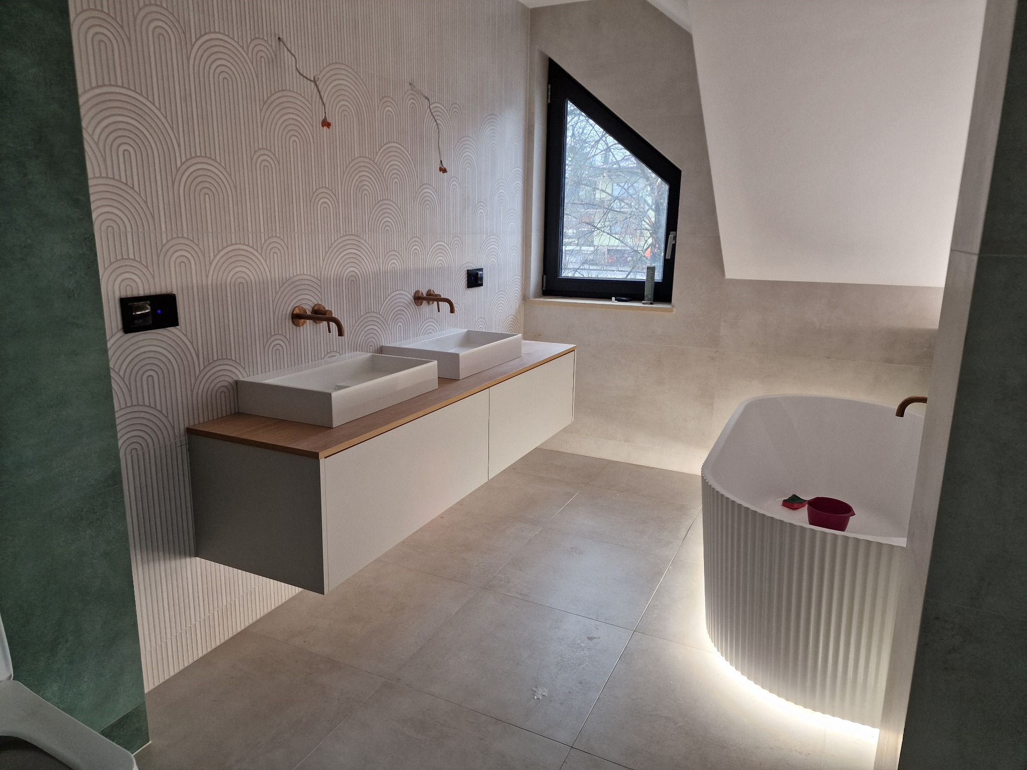 Nowoczesna łazienka z dwoma umywalkami nablatowymi na szafce z drewnianym blatem, dekoracyjną ścianą z wzorem łuków, asymetrycznym oknem i wolnostojącą wanną z podświetleniem LED u dołu.