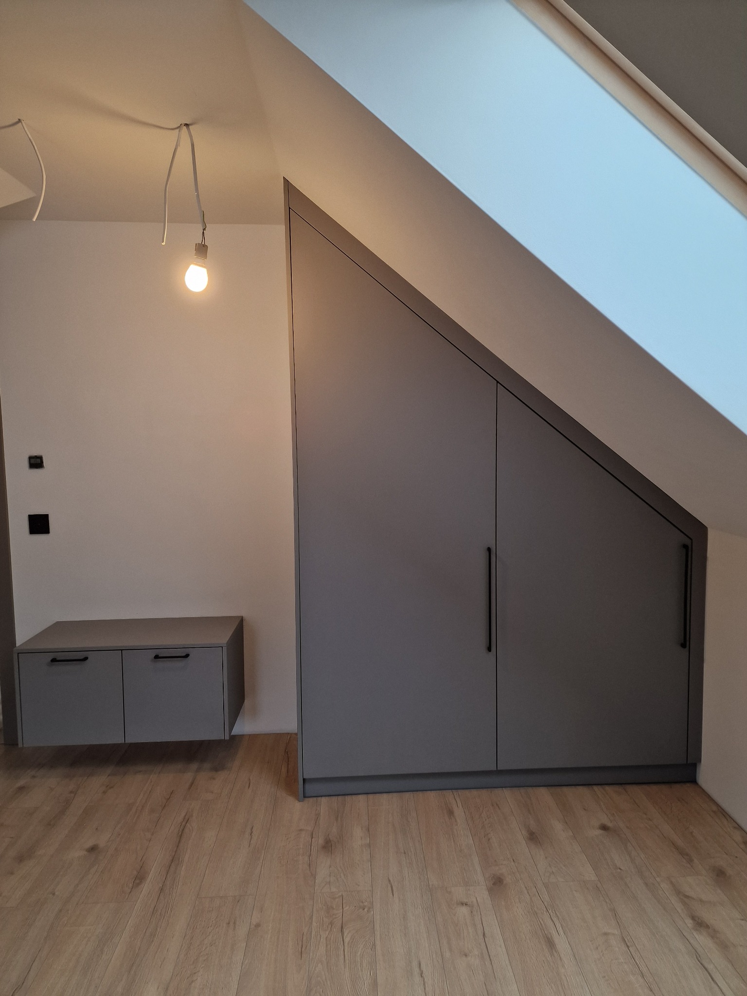 Szara zabudowa wnęki pod skosem na poddaszu, z szafką wiszącą i widoczną żarówką na kablu.