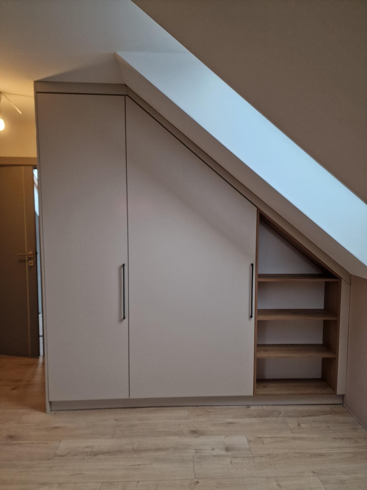 Szafa wnękowa z jasnymi frontami i czarnymi, podłużnymi uchwytami, dopasowana do skosu poddasza, z otwartymi półkami z jasnego drewna po prawej stronie.